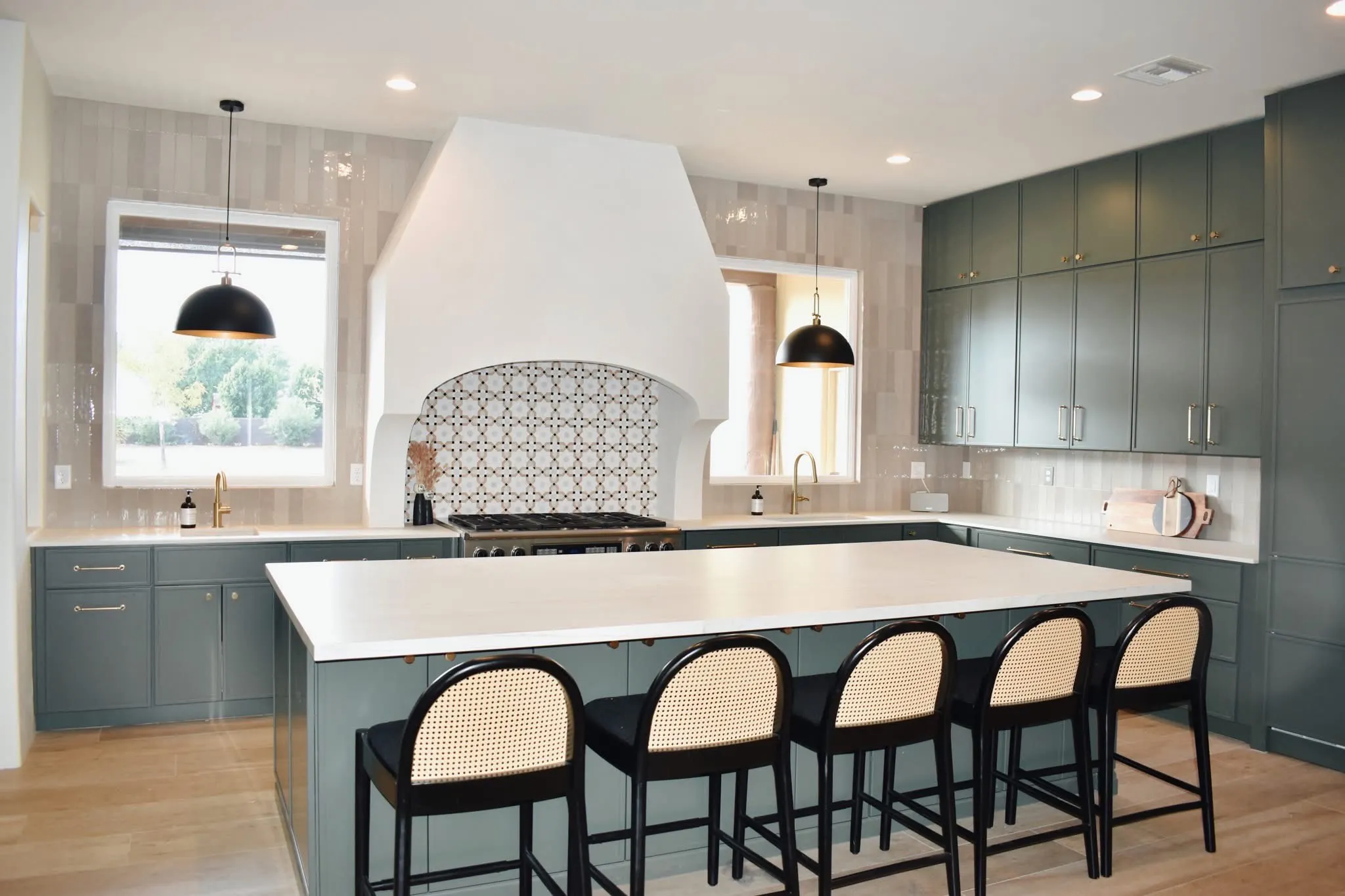 Wide view of a remodeled kitchen with green cabinetry, white island, brass hardware, and black pendant lights.
