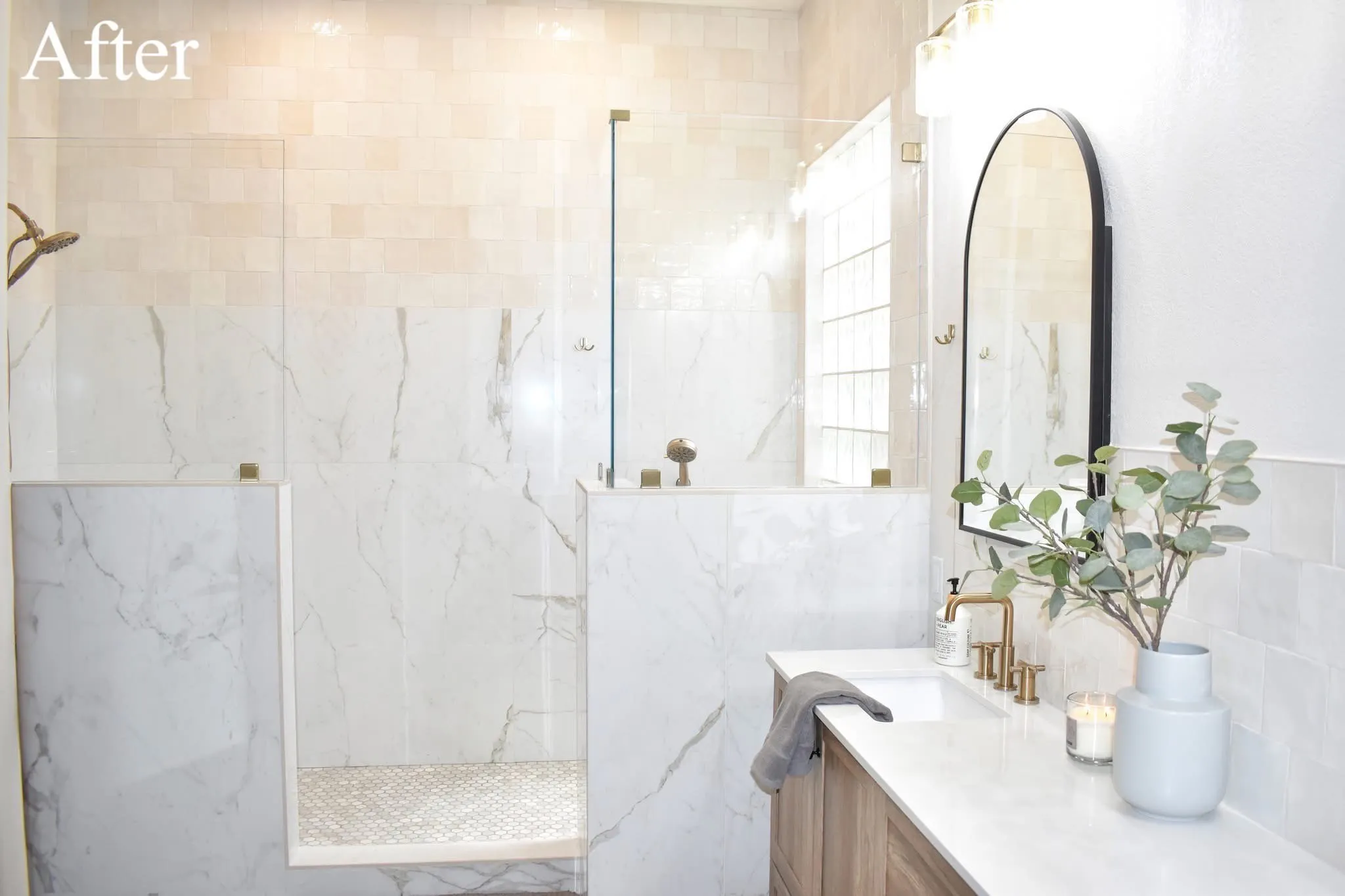 Remodeled bathroom with an open glass shower, marble-look wall panels, light wood vanity, and brass fixtures.