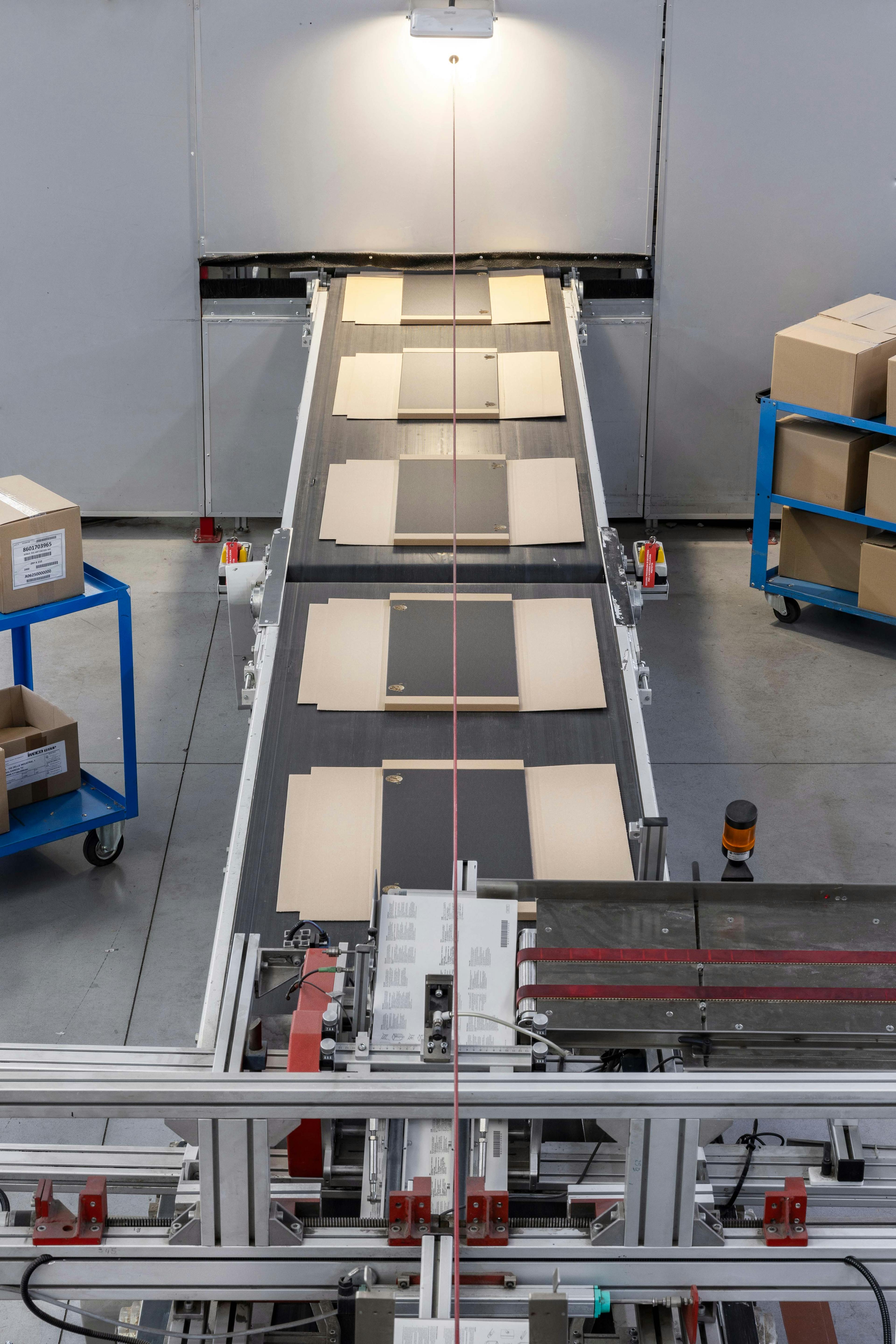 High-angle, full shot of a conveyor system for cardboard boxes, with stacked boxes in various beige and gray tones moving along a light gray metal conveyor belt.