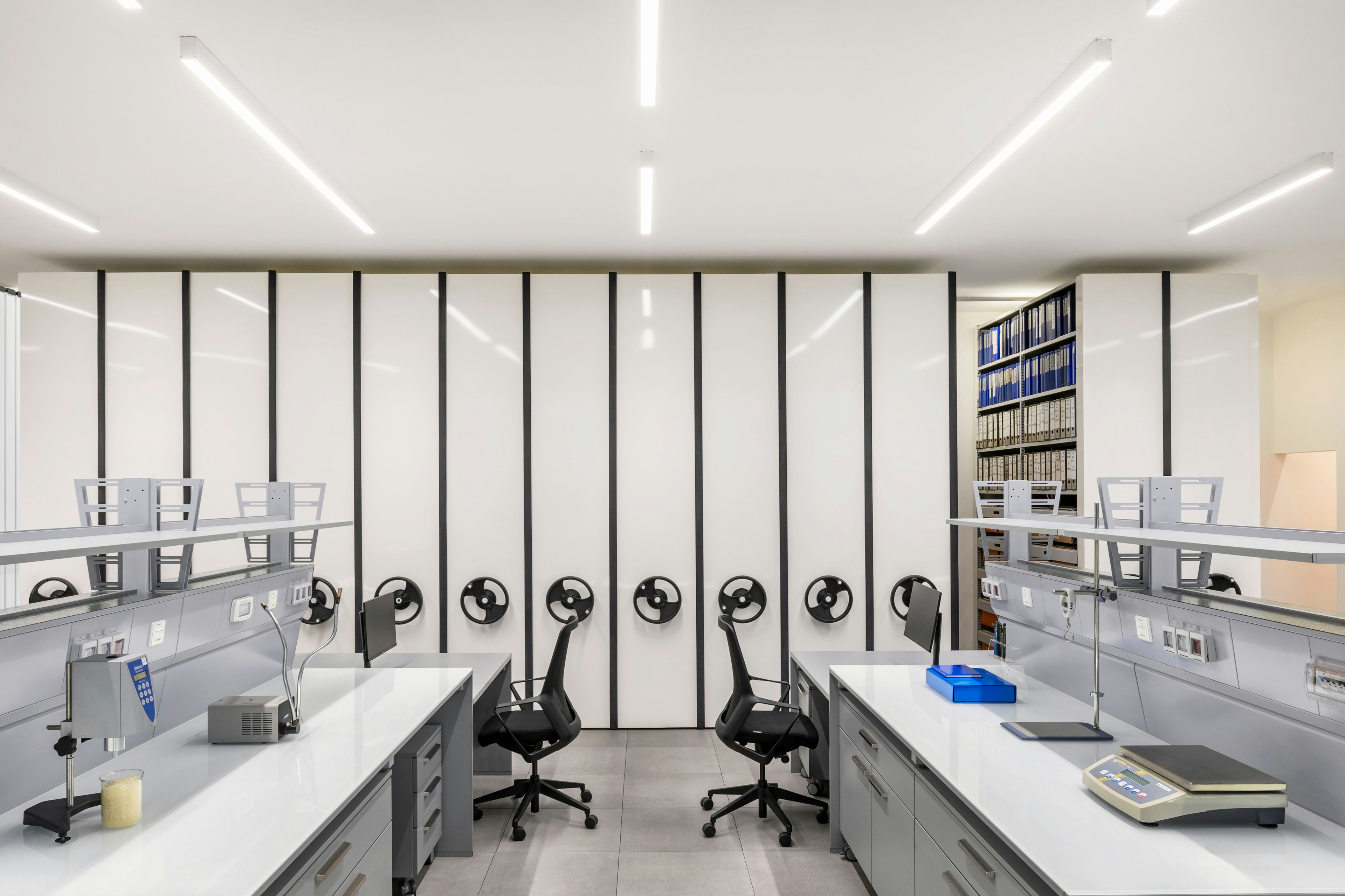 Laboratorio moderno con postazioni di lavoro ordinate, sedie ergonomiche nere e superfici di lavoro bianche; sullo sfondo armadi ad alta densità con manopole nere e scaffali pieni di raccoglitori visibili tra due pannelli aperti.