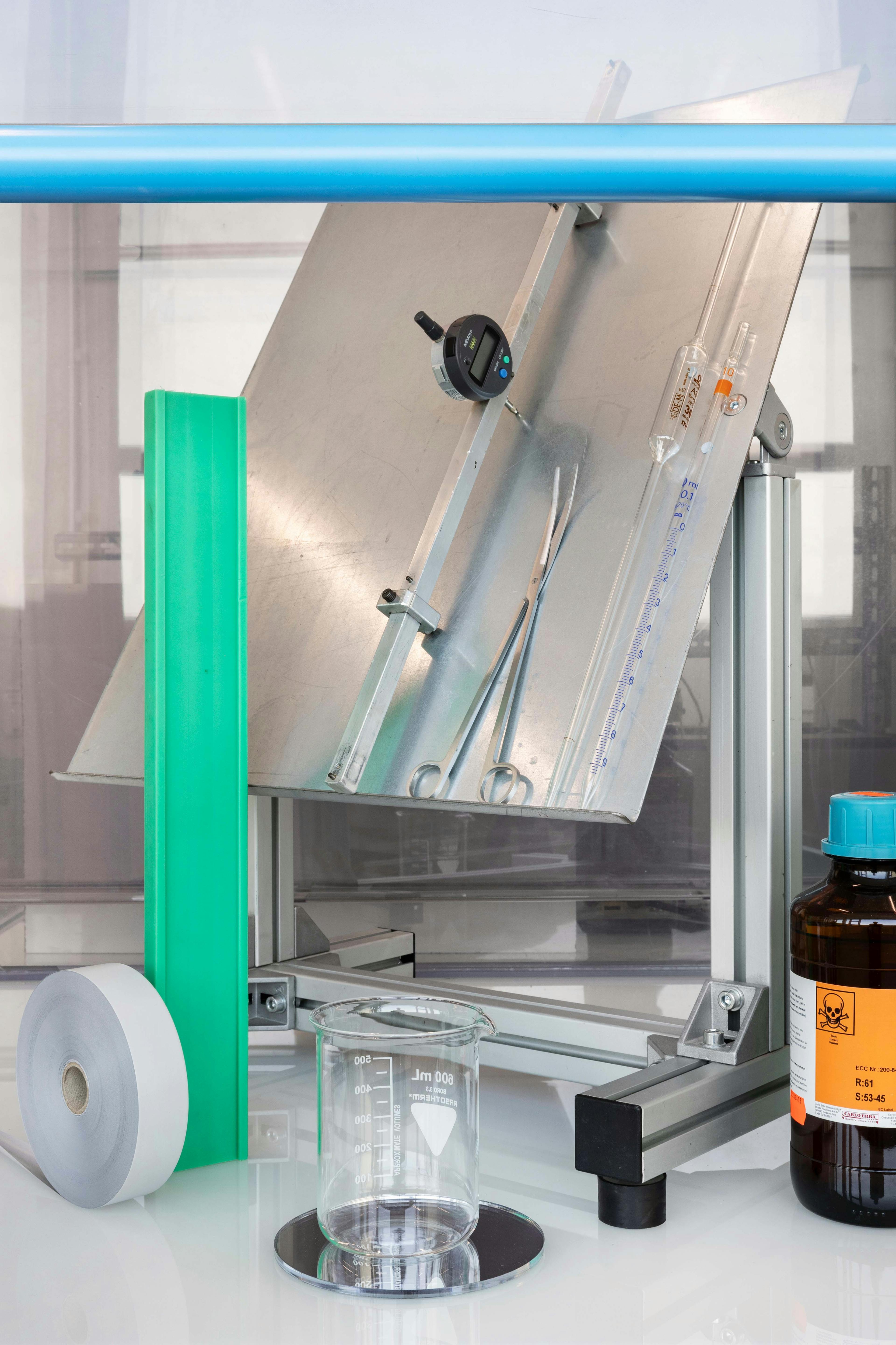 Scientific equipment inside a laboratory, including the arrangement of scientific instruments such as an inclined metal plate, a caliper, a clamp, a beaker, and brightly colored plastic strips in green and gray.