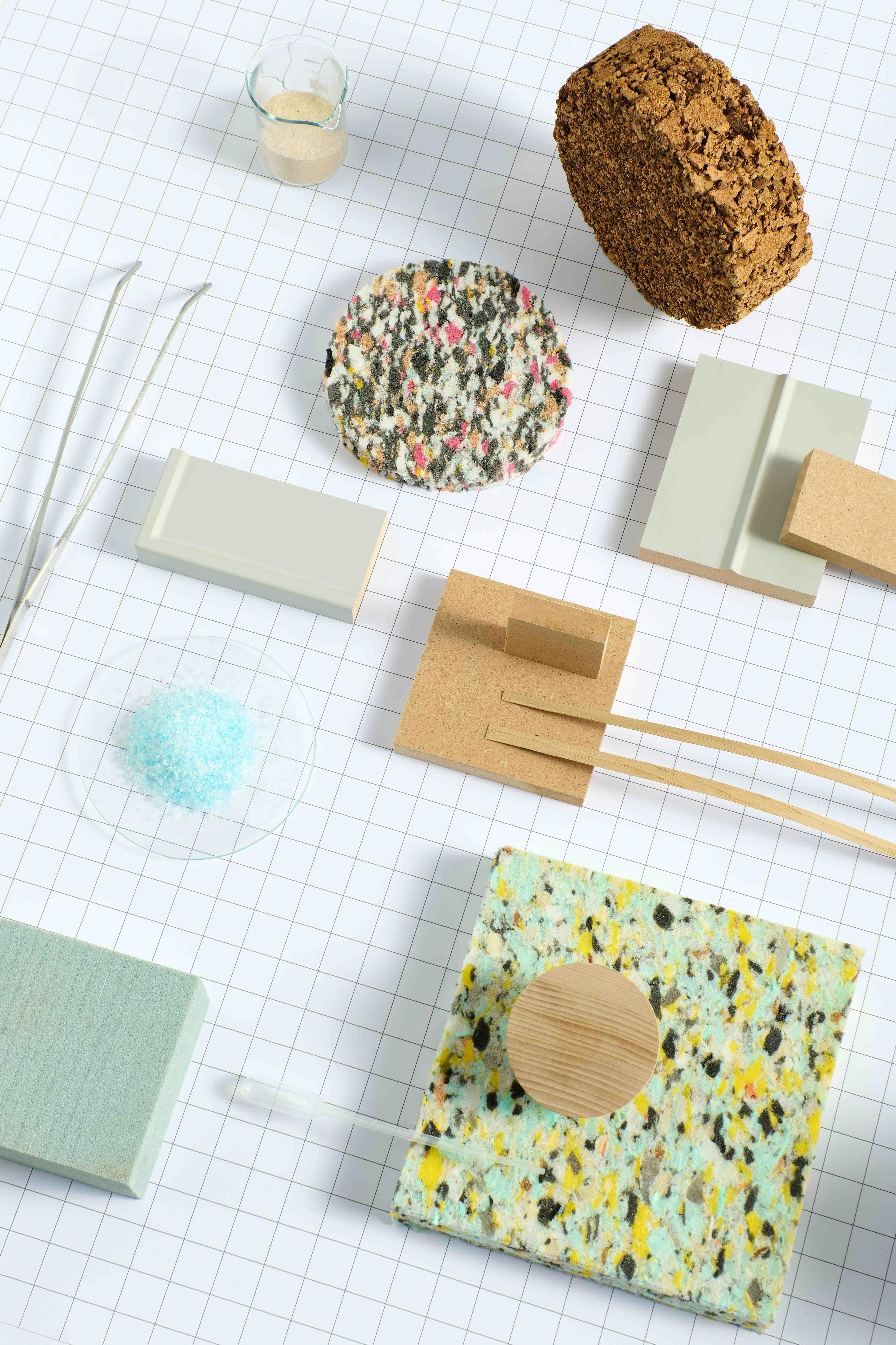 An image shows a variety of materials, objects, and samples arranged on a white grid background. Various material samples—such as blocks, fabric pieces, foam, caps, and other elements—are distributed across the space.
