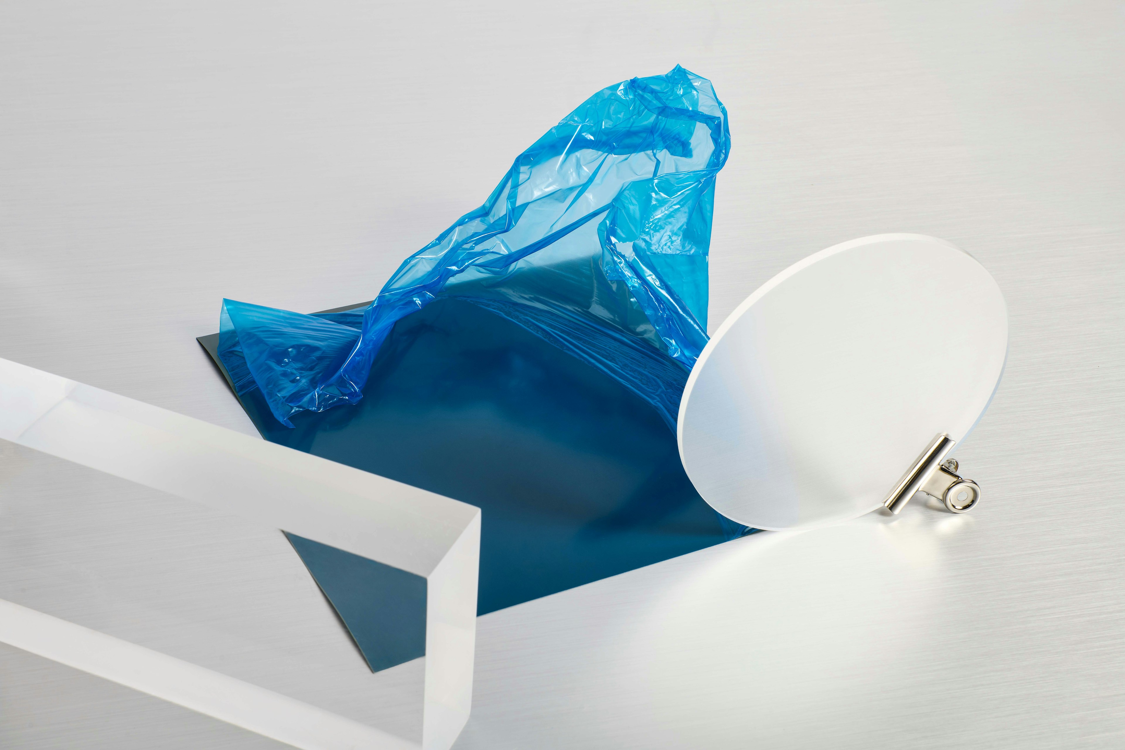 Un foglio rettangolare di materiale di colore verde acqua, liscio, ricoperto da una pellicola di plastica traslucida e spiegazzata di colore azzurro chiaro, poggia su una superficie grigio chiaro/bianca.