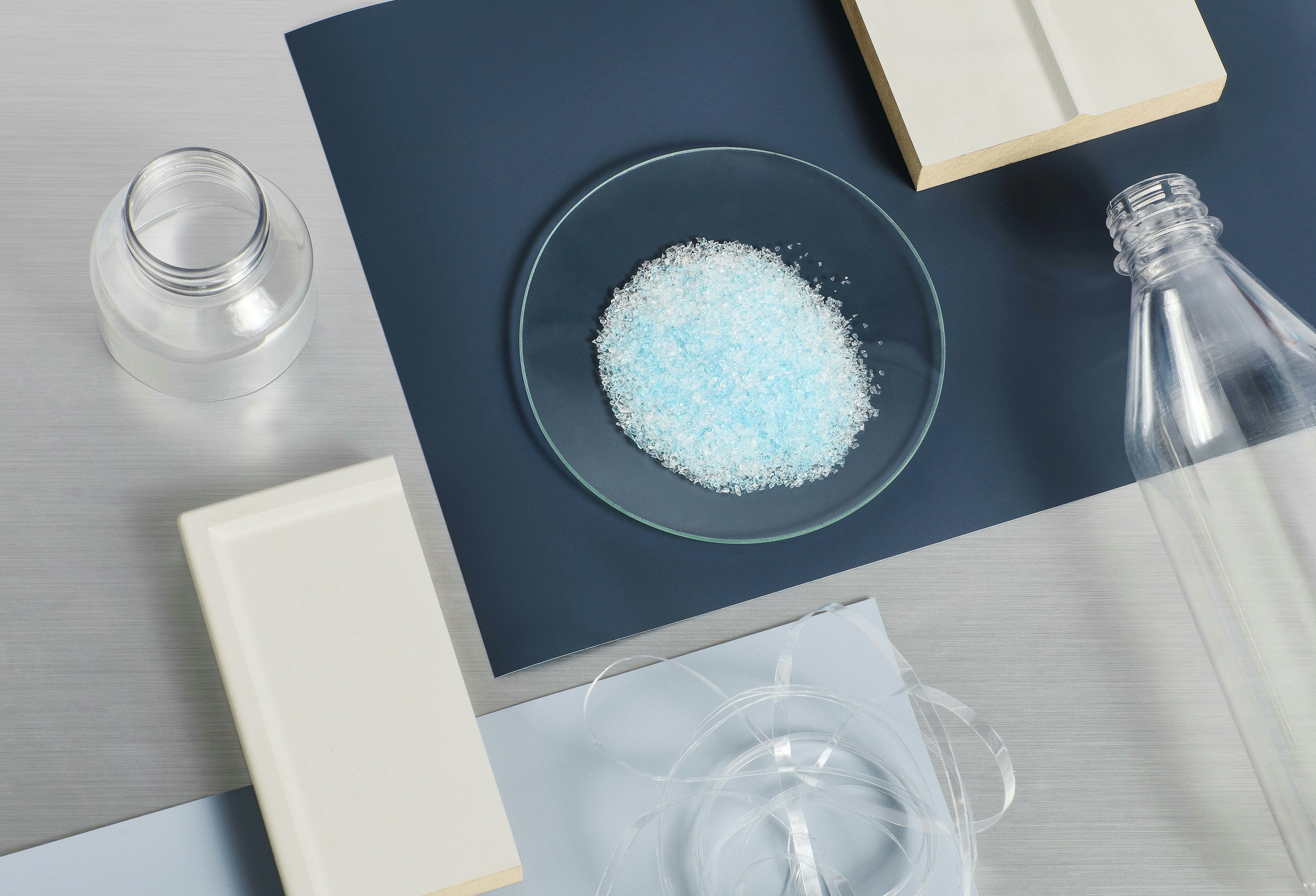 A high-angle view of various laboratory materials, including clear plastic bottles, a glass plate with light blue granular material, a white rectangular tile, and translucent plastic wrap.