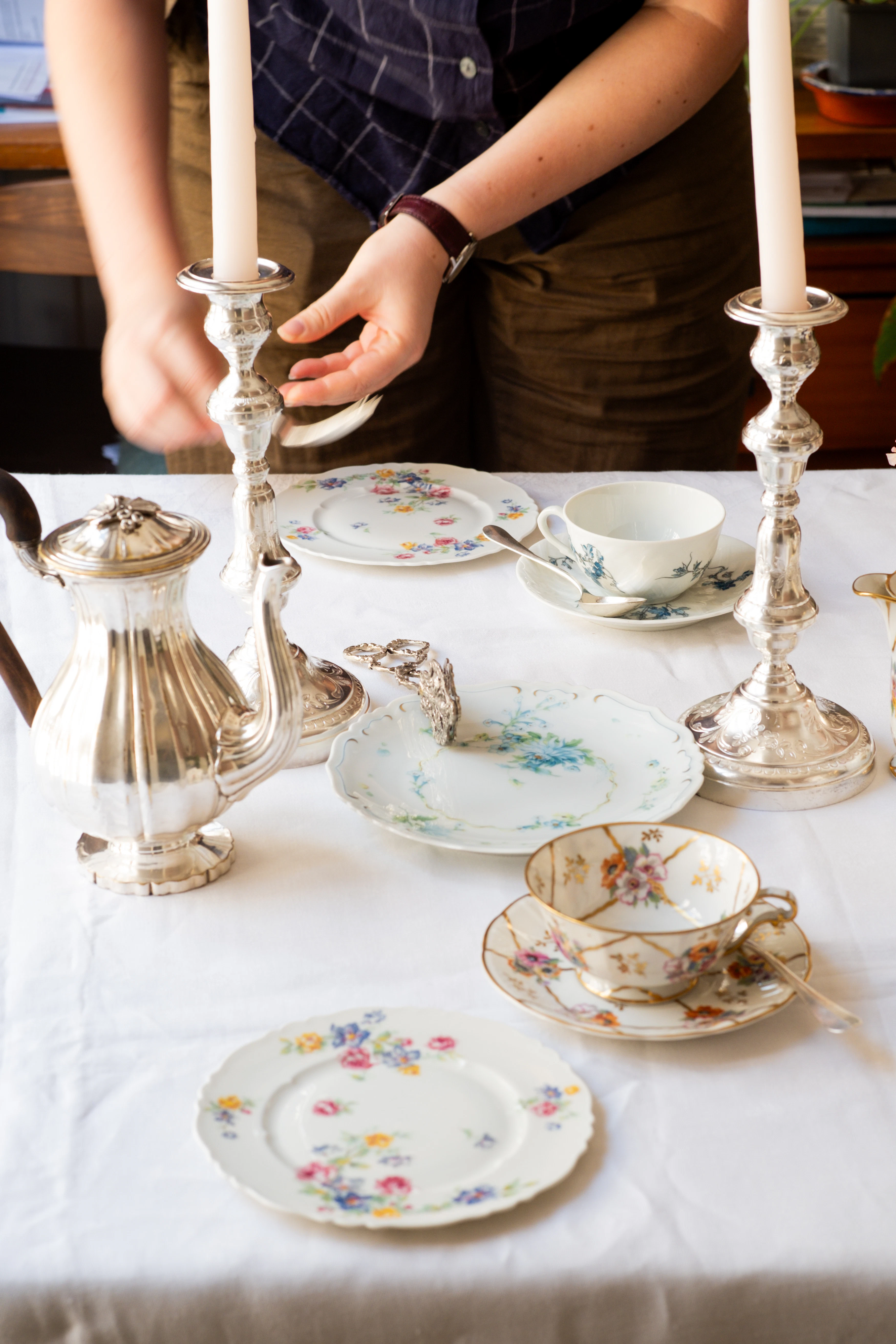 Two hands setting a table with two porcelain table settings and two silver candlestick holders with two cream candles.