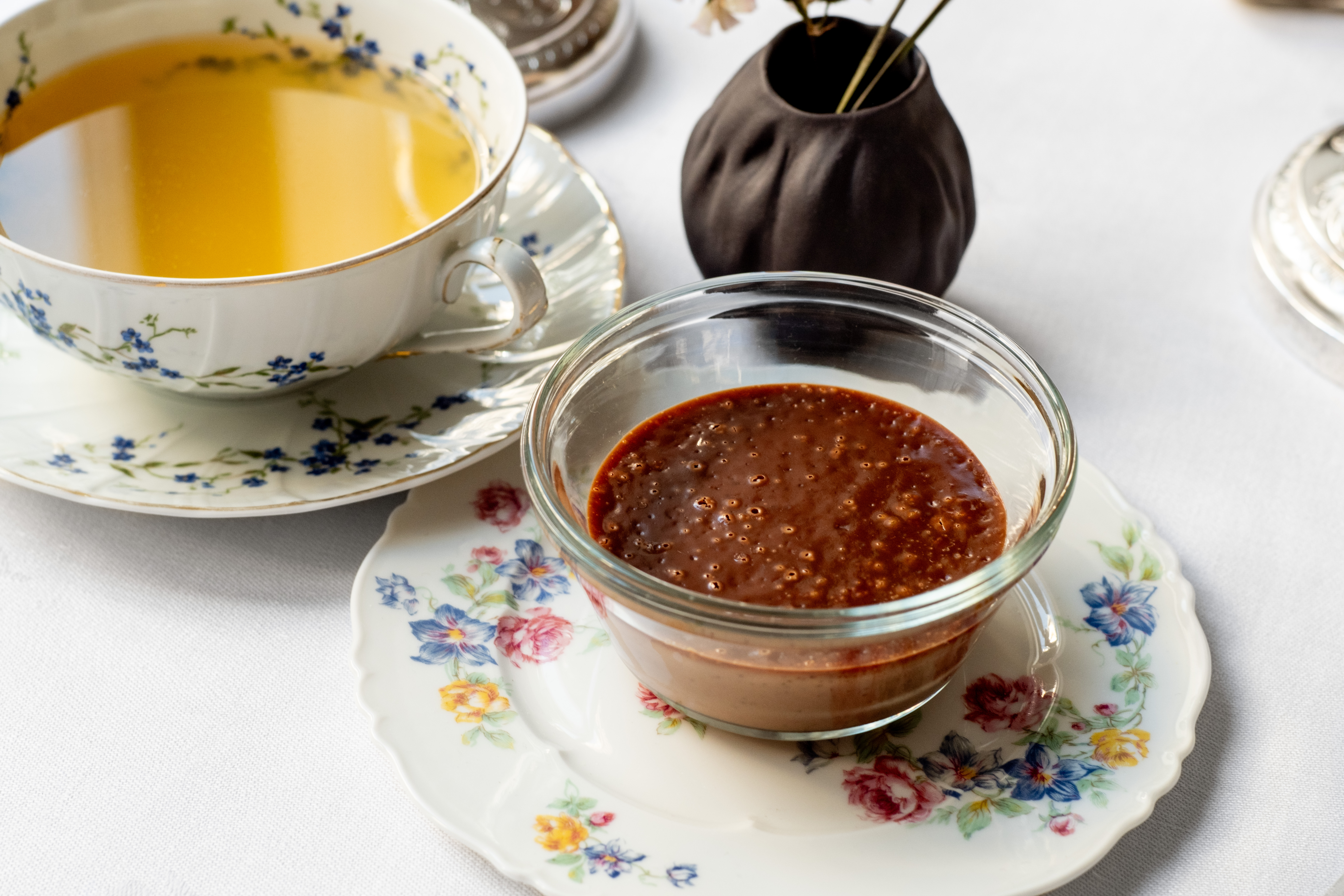A chocolate cream dessert sitting on a floral print plate. In the background is a pale yellow tea in a floral cup on a saucer.