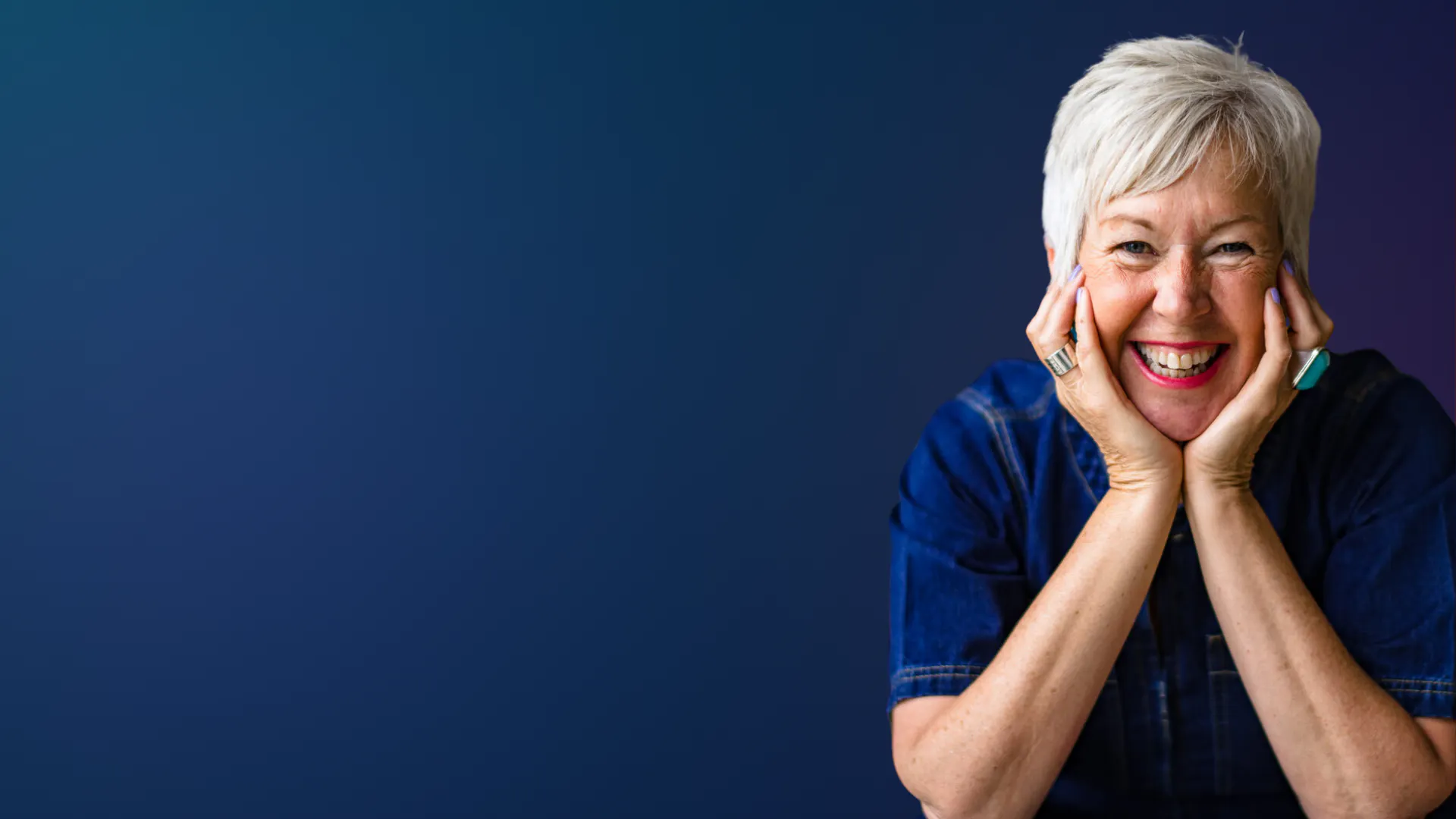Smiling woman with short grey hair resting her chin on her hands against a dark blue background