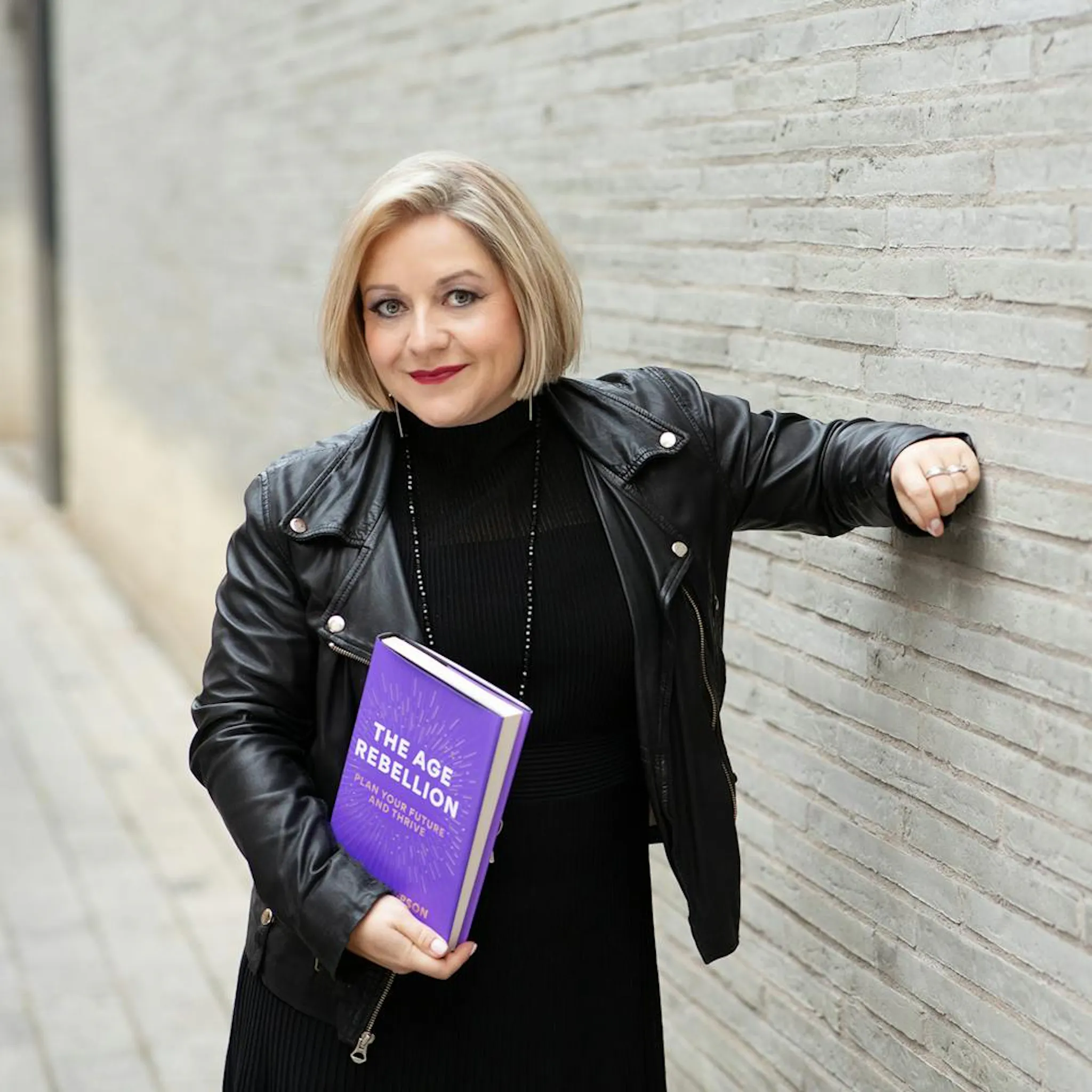 Lyndsey Leaning against a wall wearing a black leather jacket holding a copy of the book