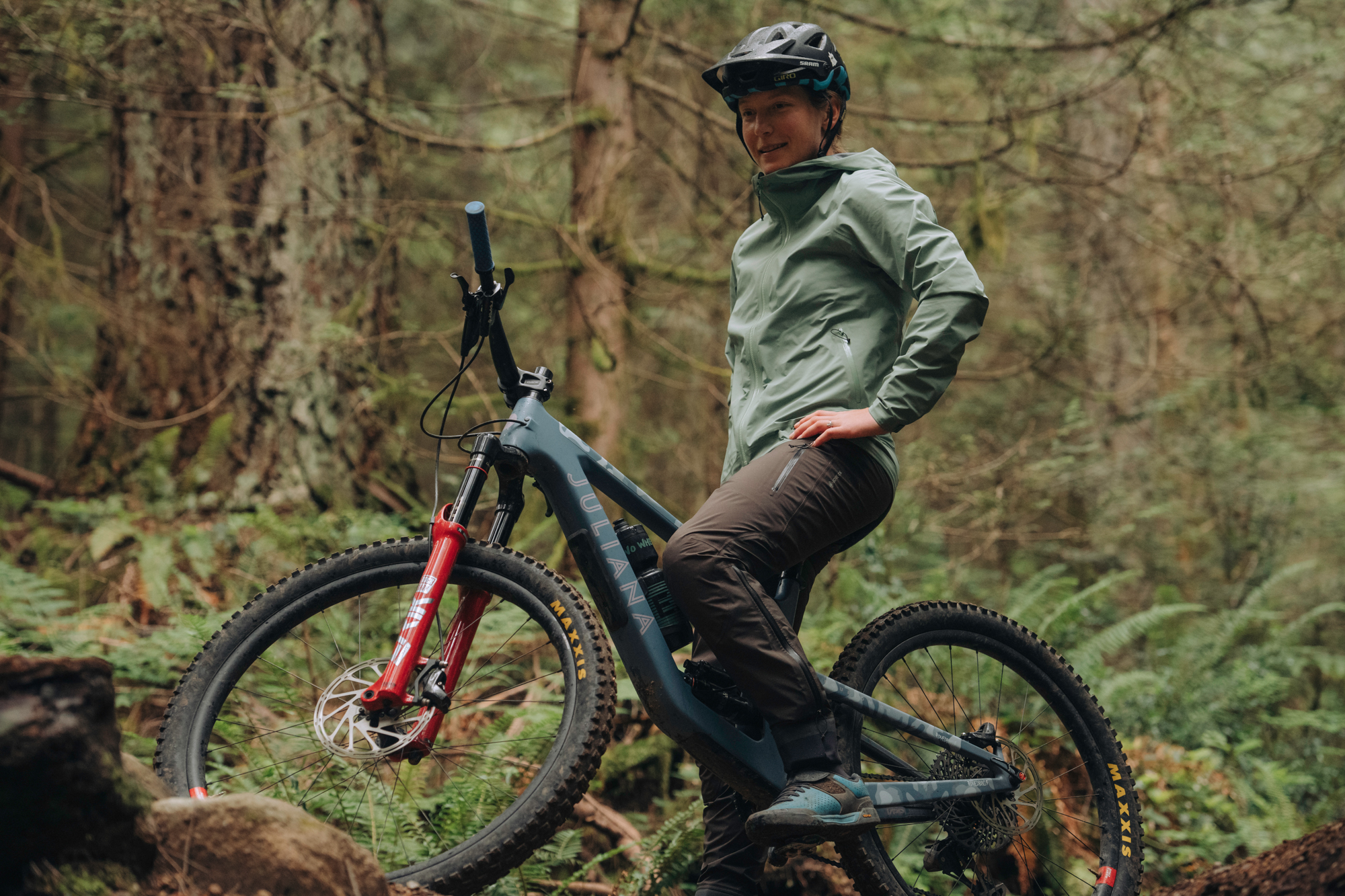 Rider in a pale green shell standing over a mountain bike in wet forest