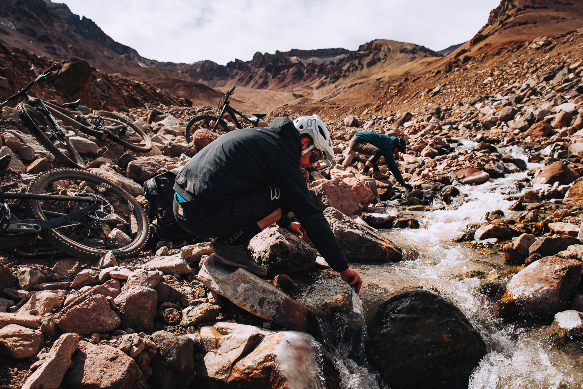 Rider filling bottles from a meltwater stream beside bikes on the rocks