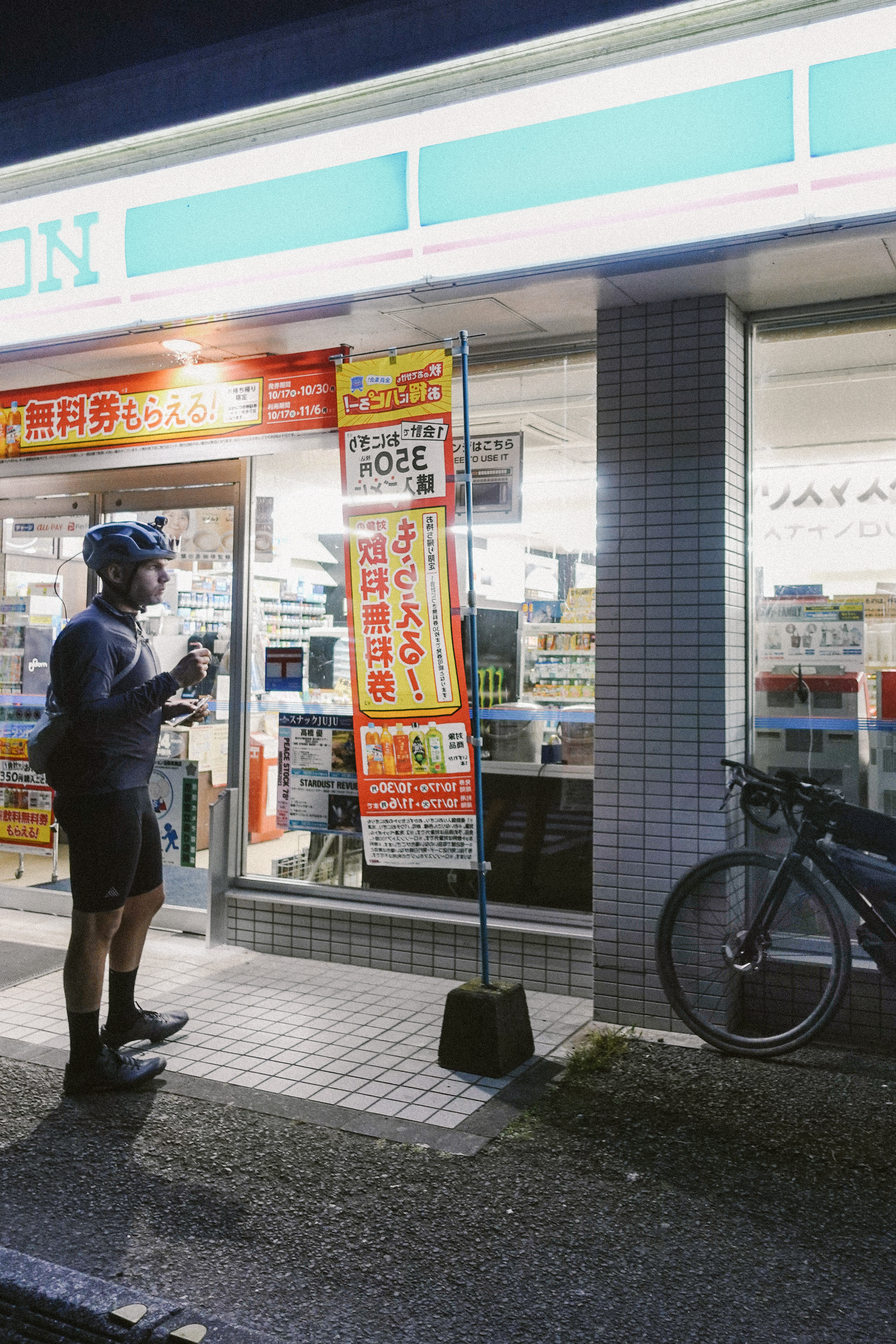 Rider eating outside a lit convenience store at night, bike parked alongside