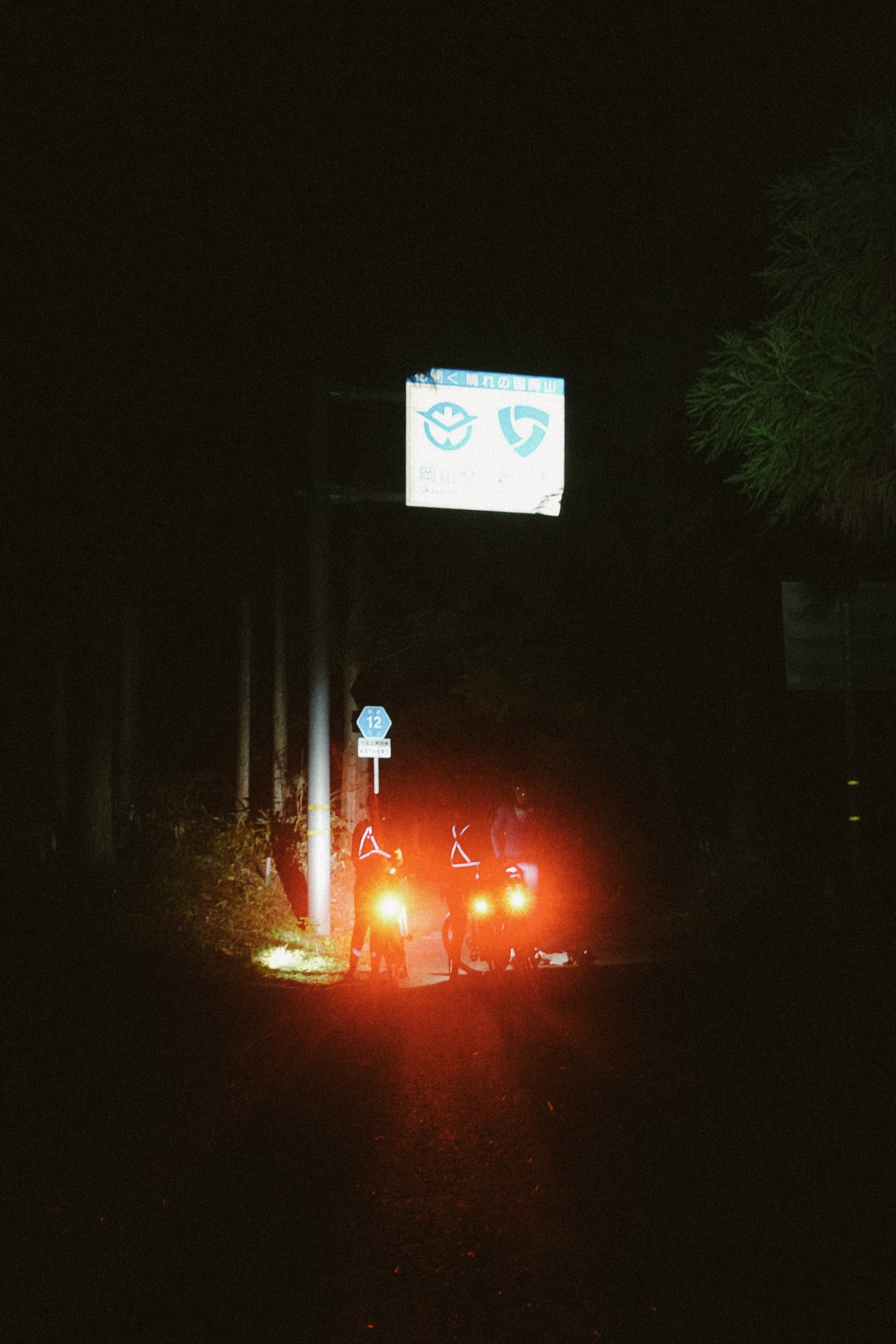 Riders stopped at night beside a route sign, lit red by their rear lights