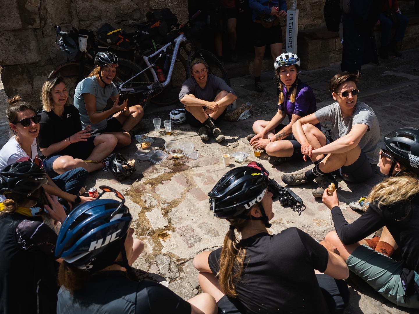 Riders sitting on the ground in a shaded street with food and drinks, helmets off