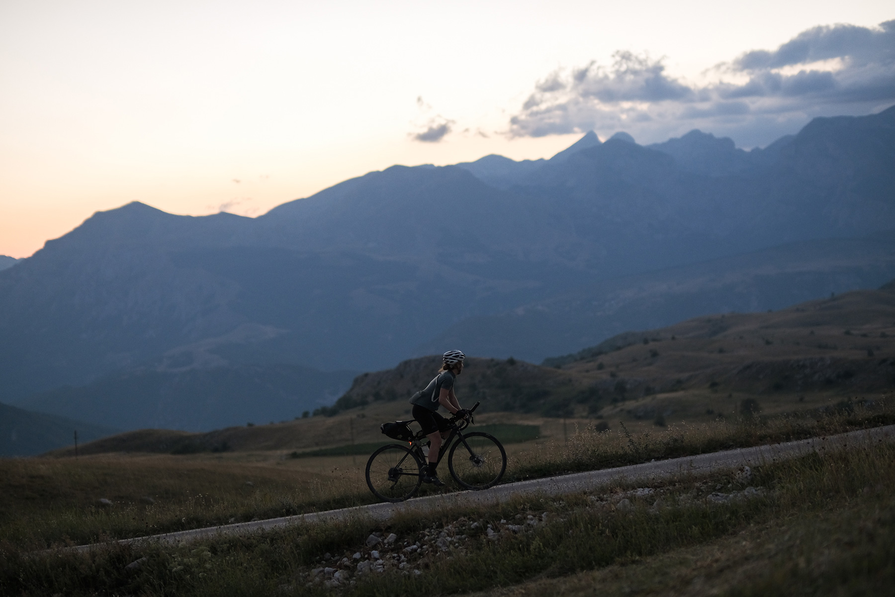 Fiona Kolbinger rides a lone road across a high plateau at dusk, mountain ridges behind her