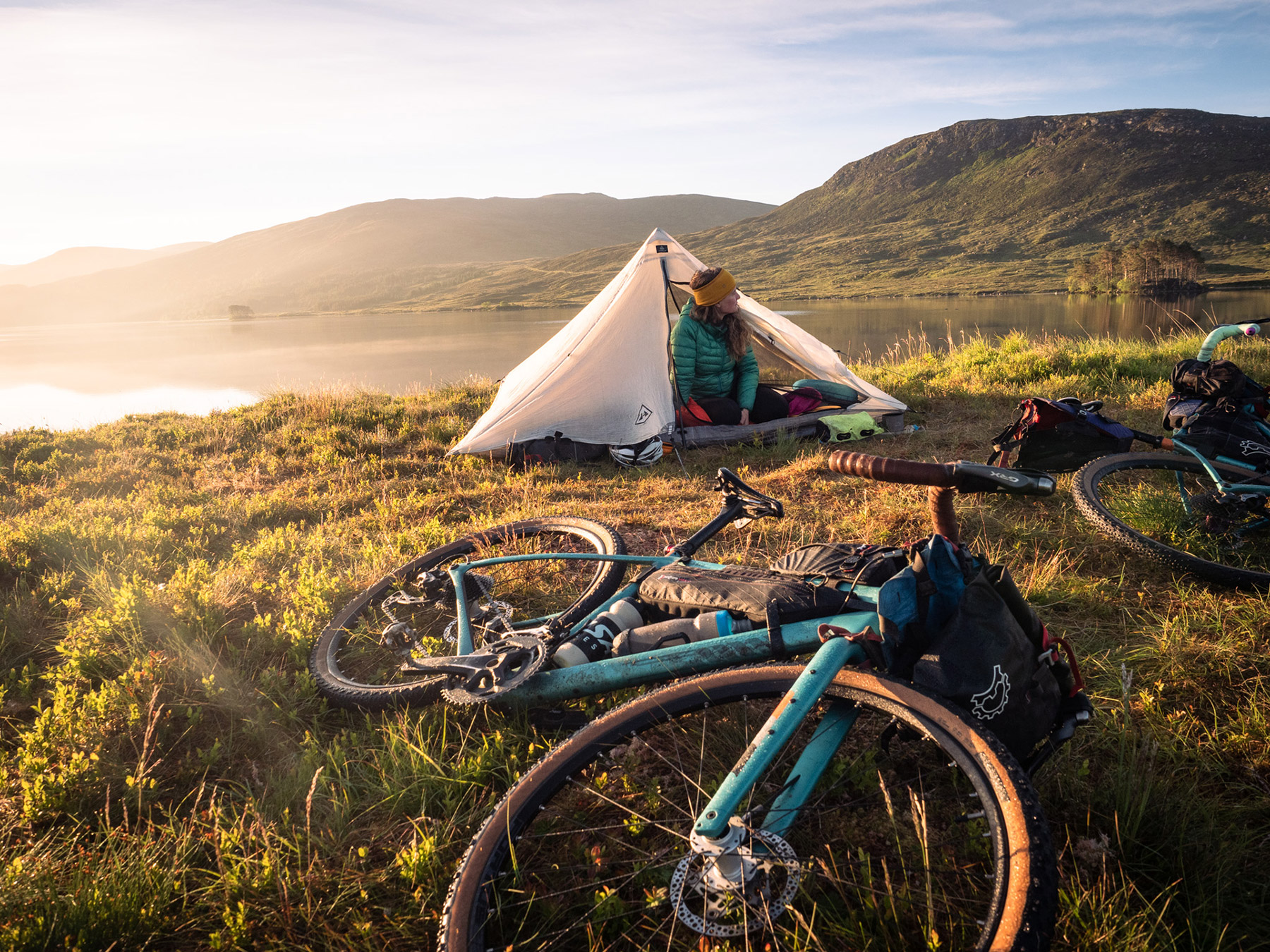 Rider sitting in a pyramid tent beside a loch at sunrise, two loaded bikes on the grass