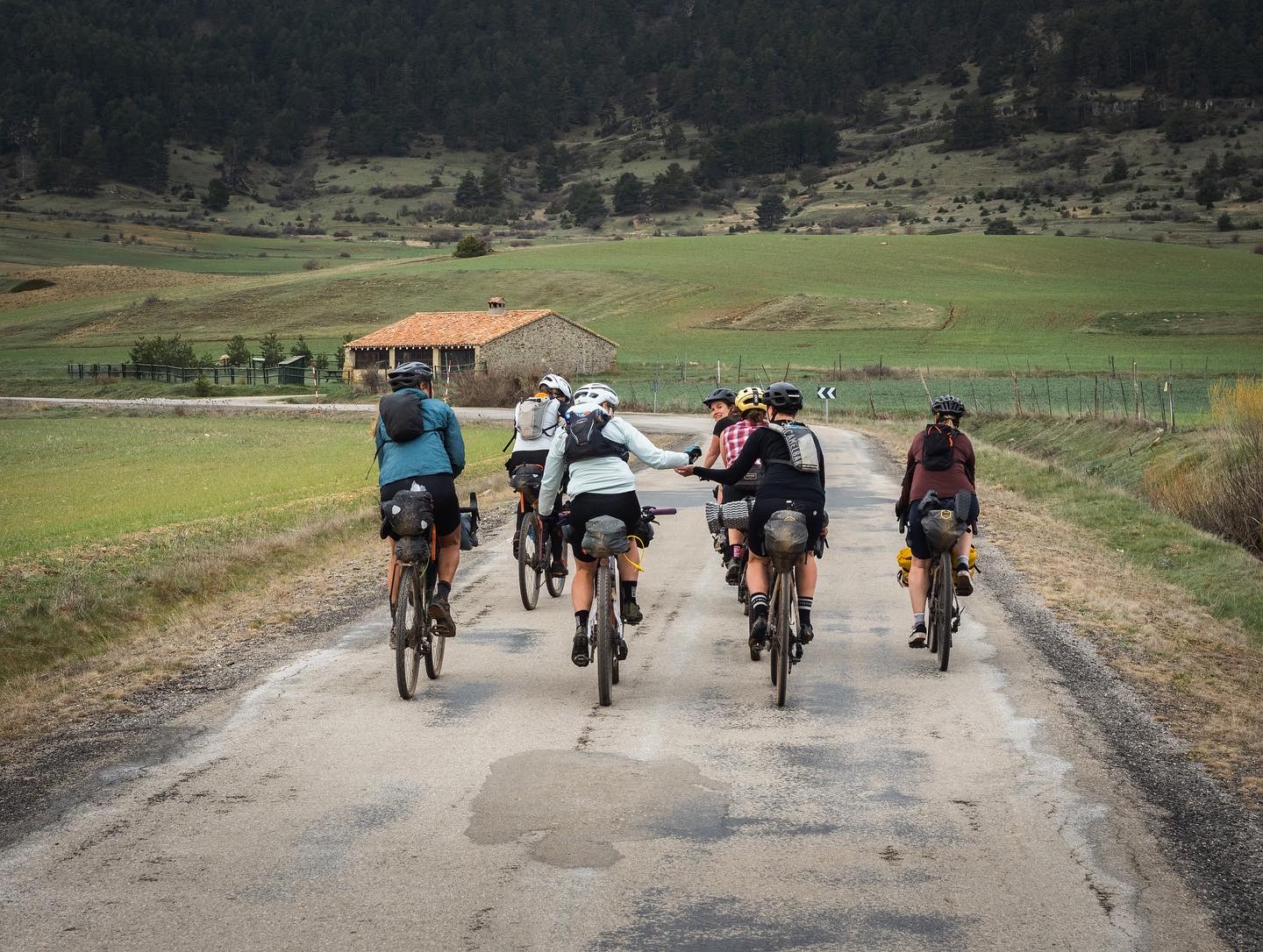 Group of loaded riders on a road through green fields with a stone hut ahead