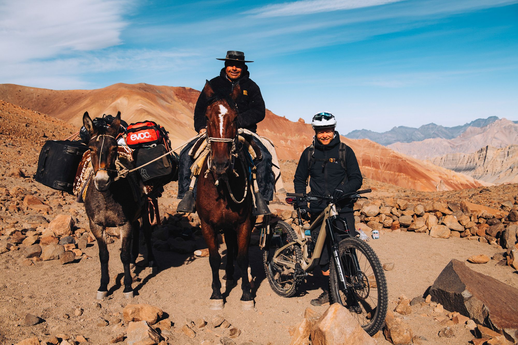Arriero on horseback with a loaded mule beside a rider and bike on red gravel