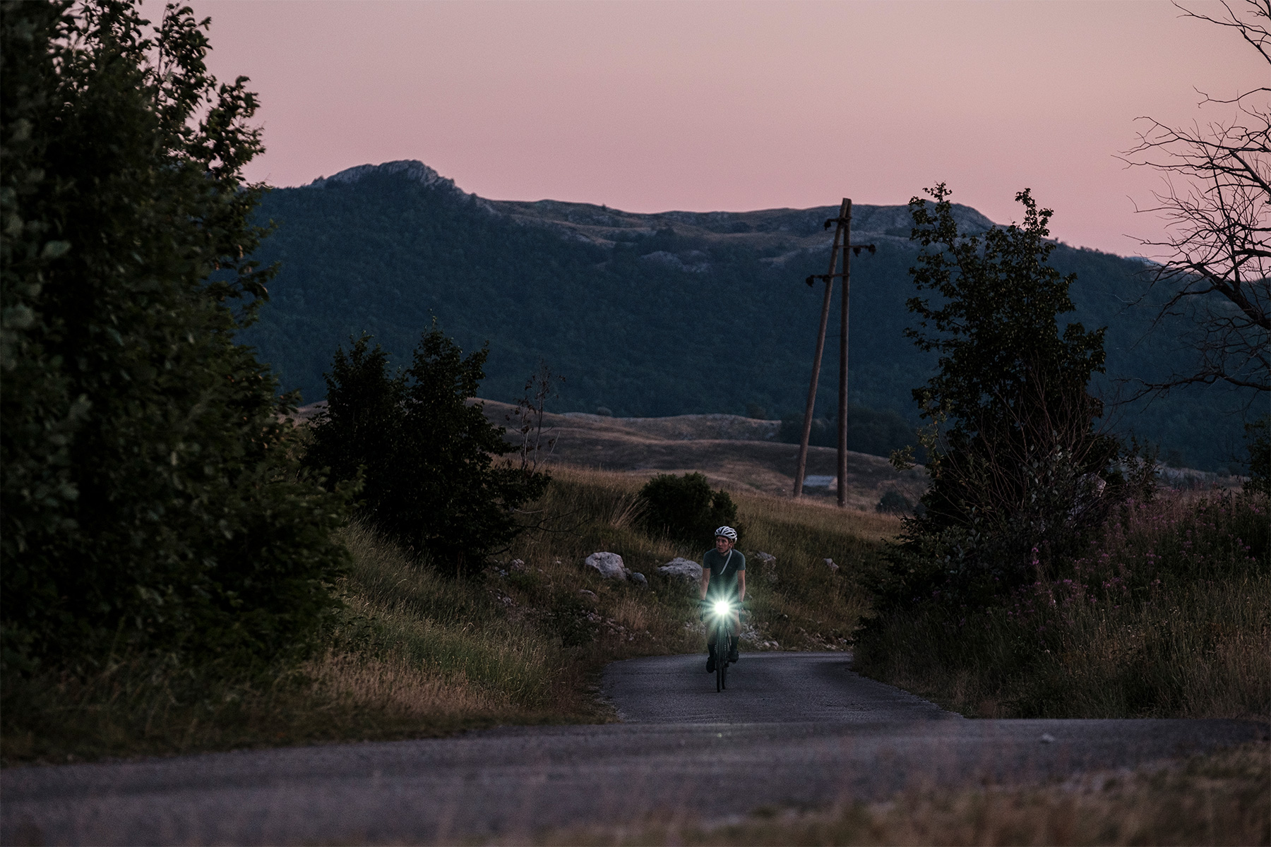 A rider comes up a narrow lane at dusk with a headlight on, pink sky over limestone hills