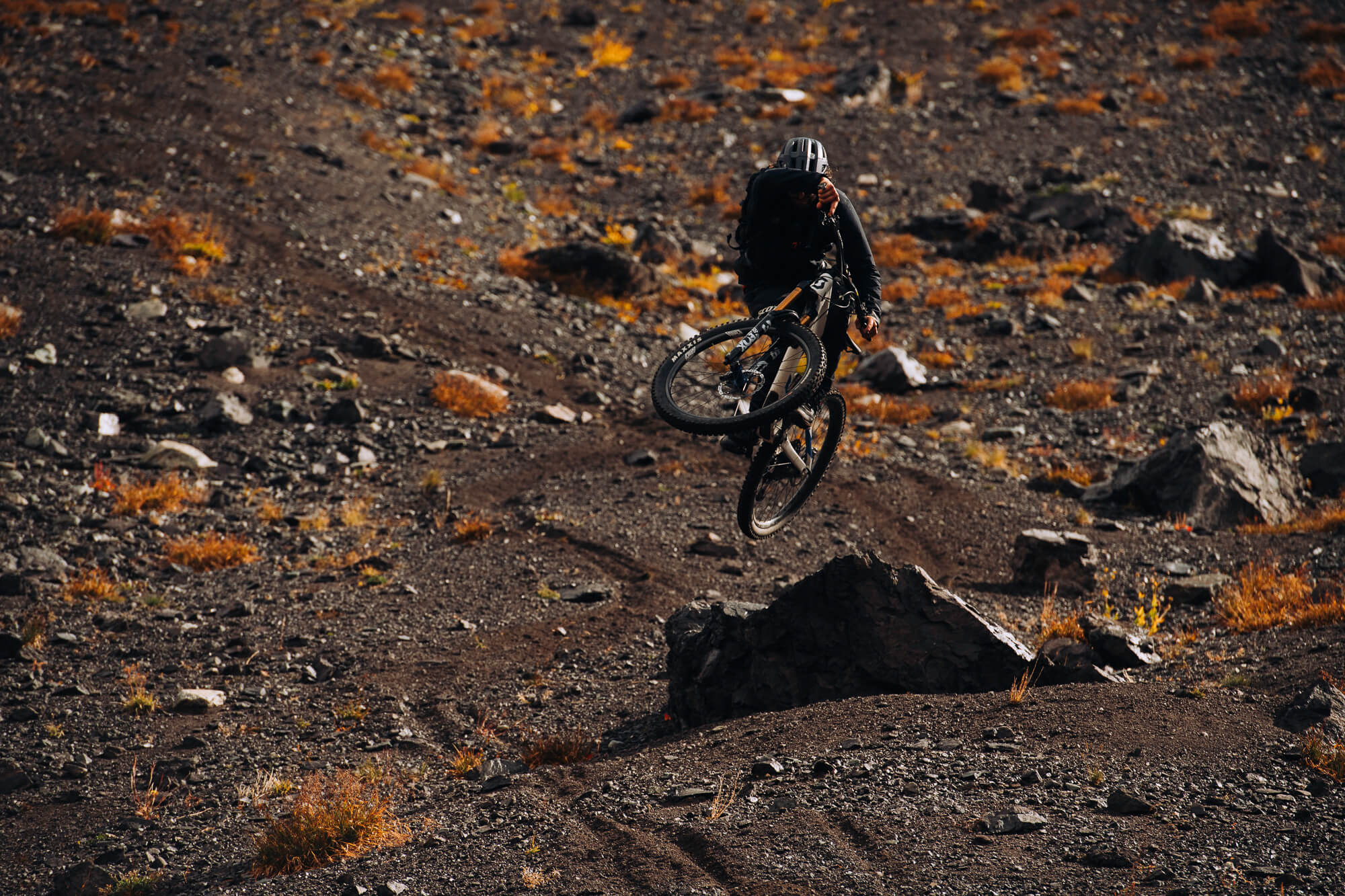 Rider launching off a rock on a loose slope dotted with autumn brush