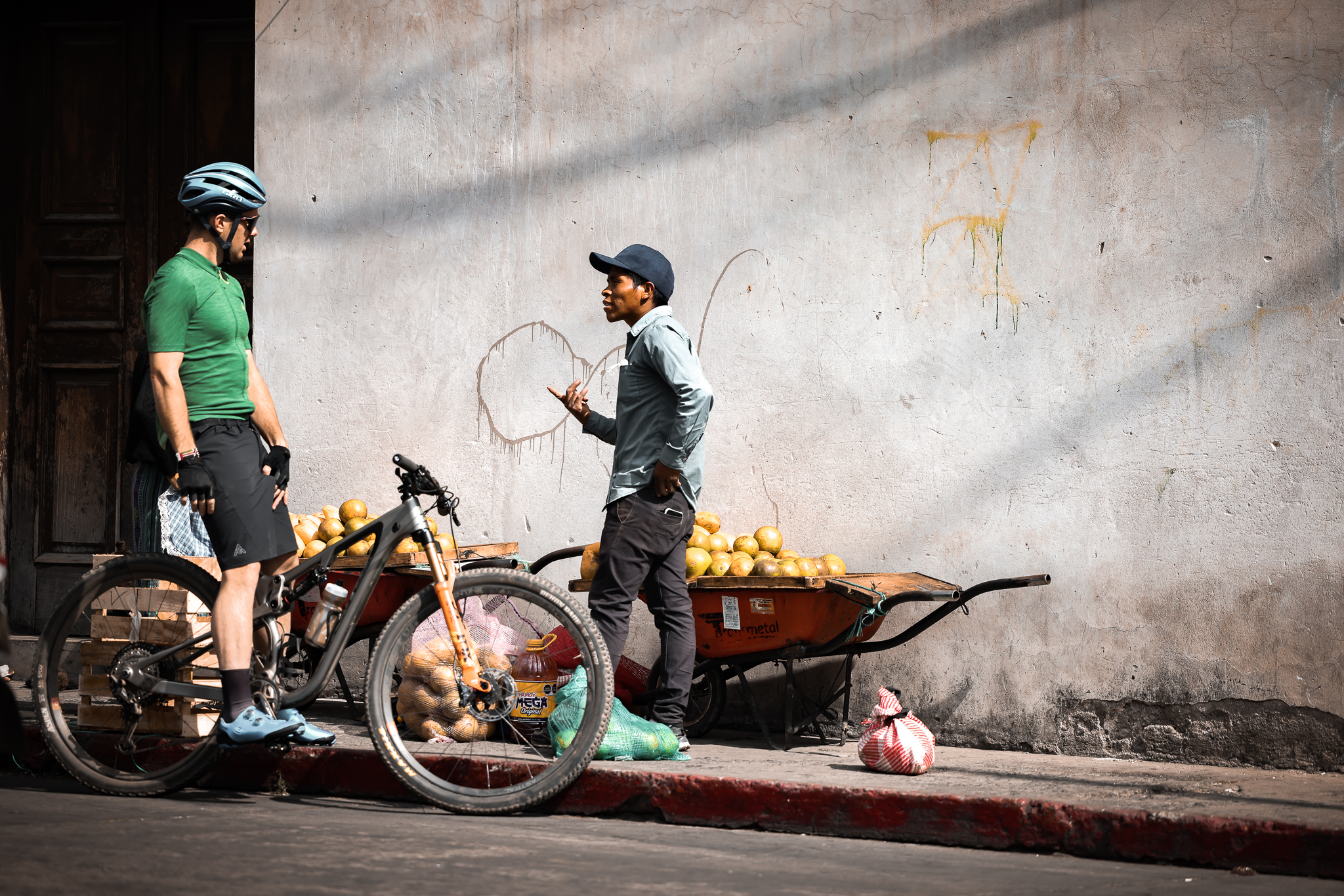 Rider with a mountain bike talking to a vendor beside a wheelbarrow of melons