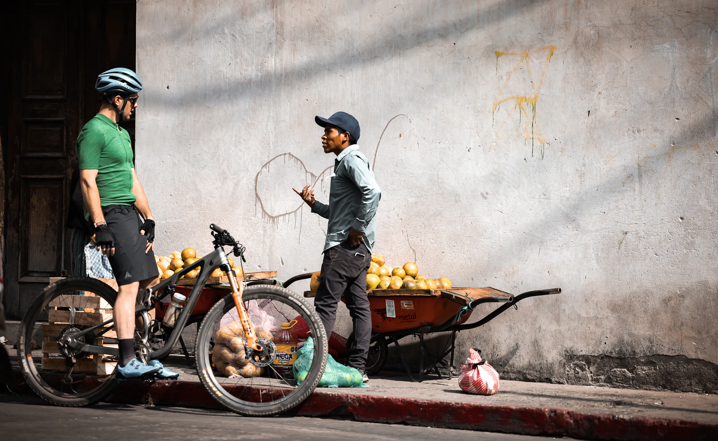 Rider with a mountain bike talking to a vendor beside a wheelbarrow of melons