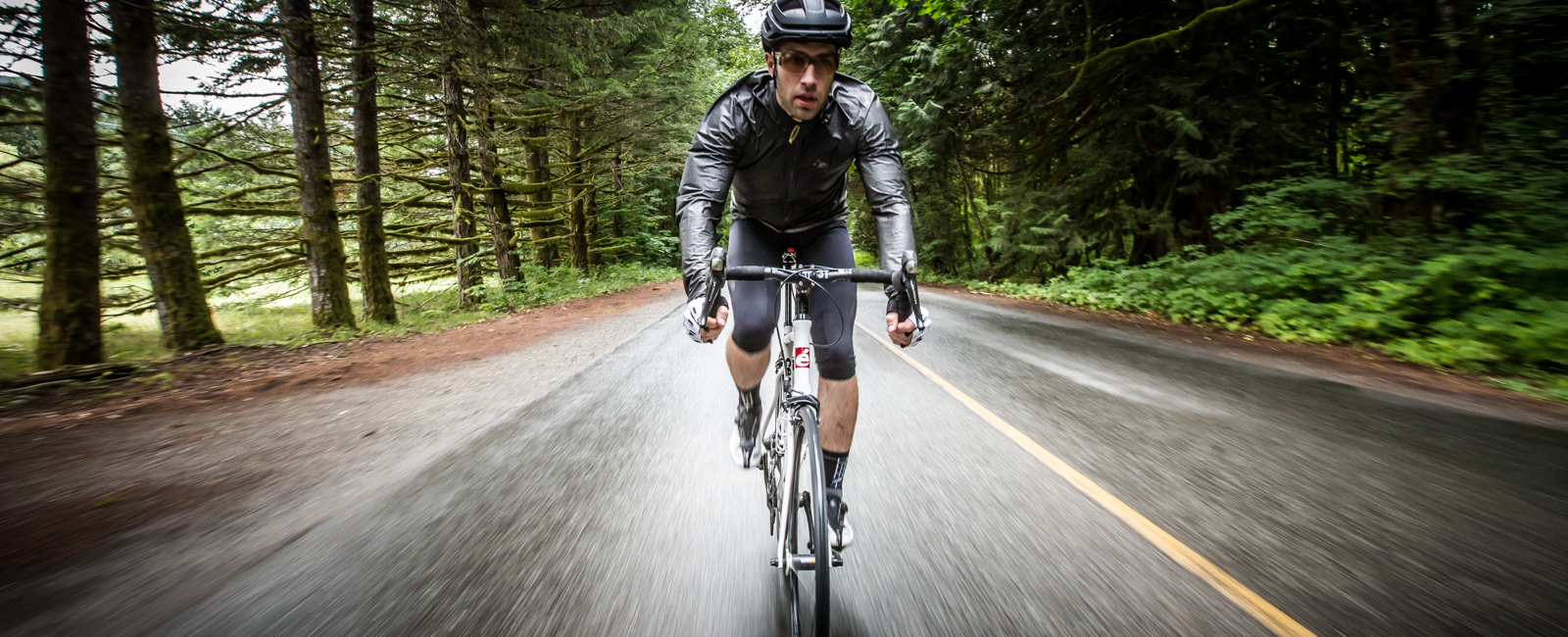 Rider in a black Oro jacket riding toward the camera on a wet road through conifer forest