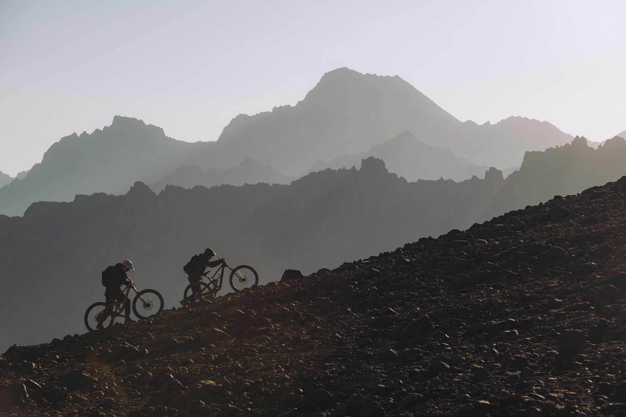 Two riders silhouetted pushing bikes up a dark scree ridge at sunrise