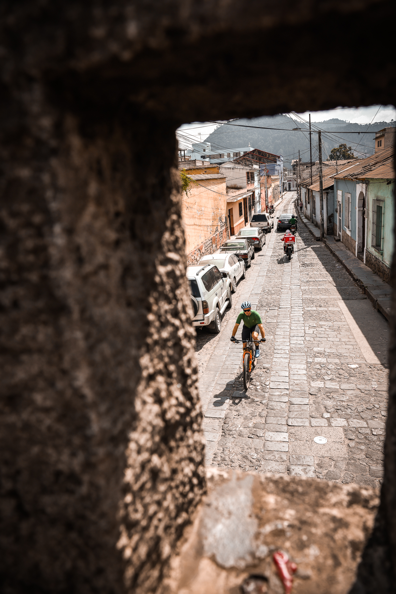 Rider climbing a narrow cobbled street past parked cars and a motorbike