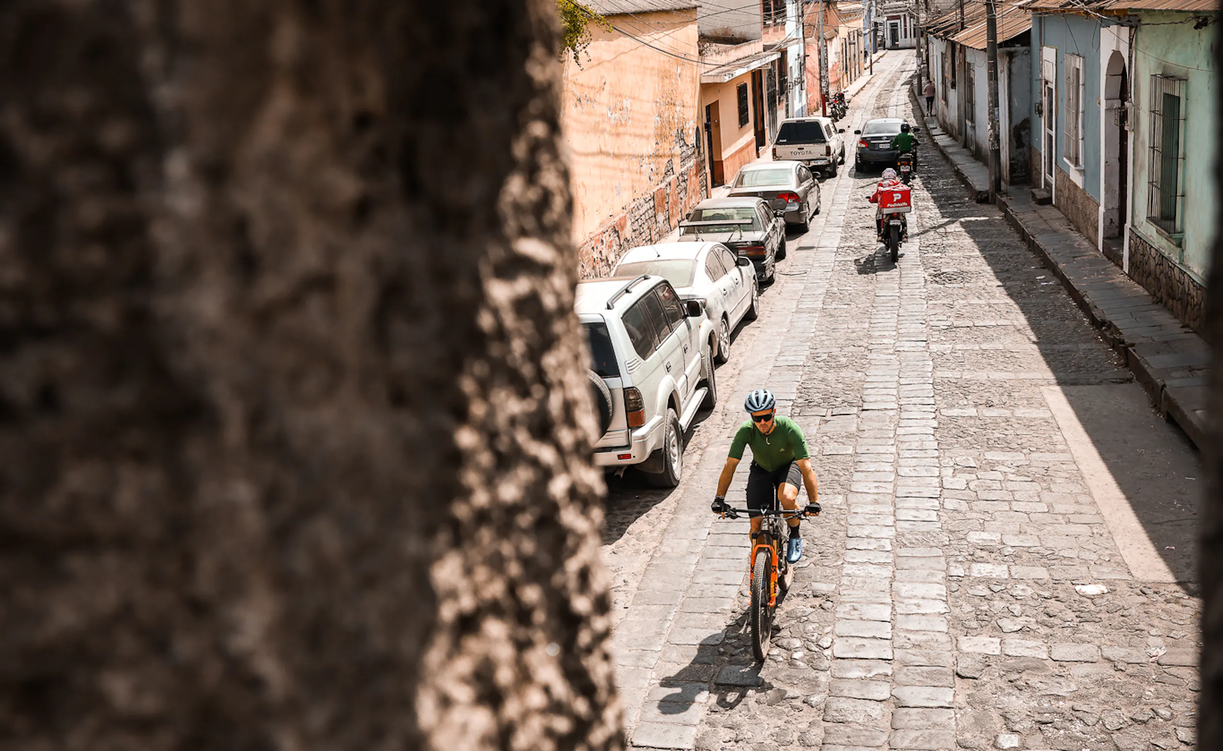 Rider climbing a narrow cobbled street past parked cars and a motorbike