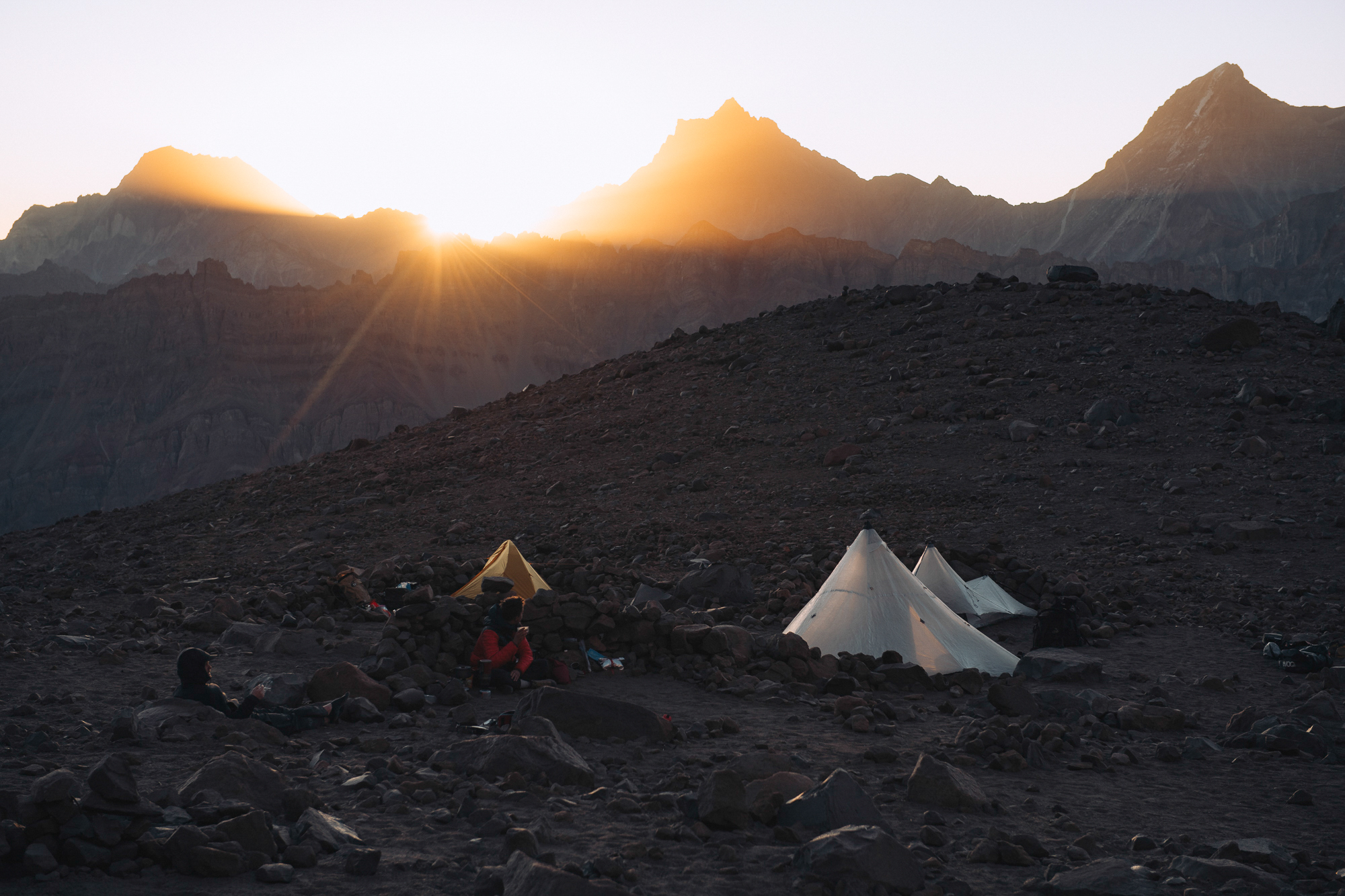 Two tents at a rocky basecamp with the sun rising over a peak behind