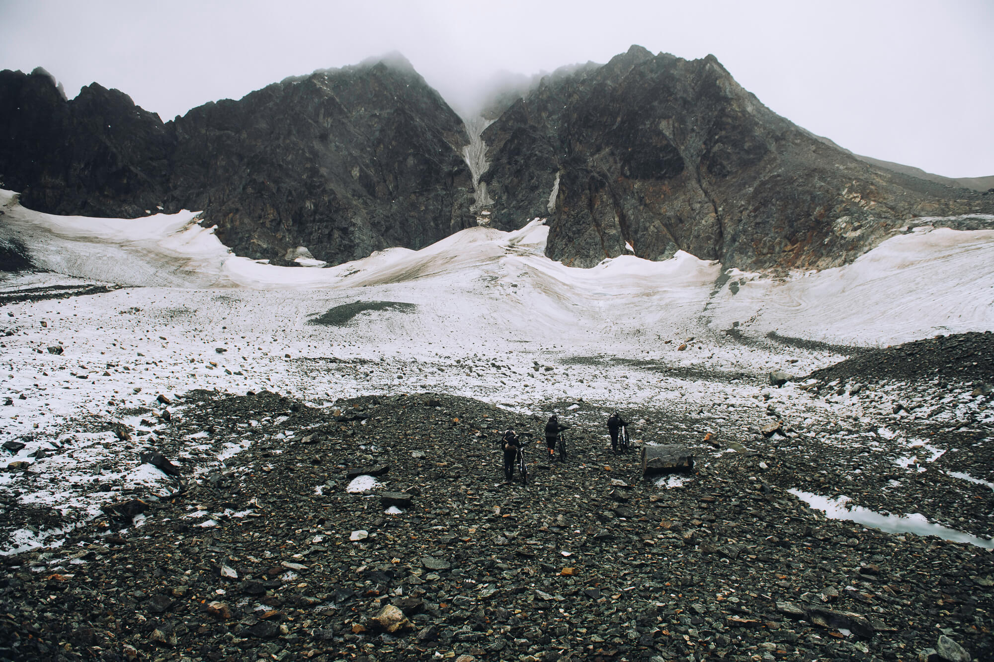 Three riders pushing bikes across rocky moraine below a snowfield