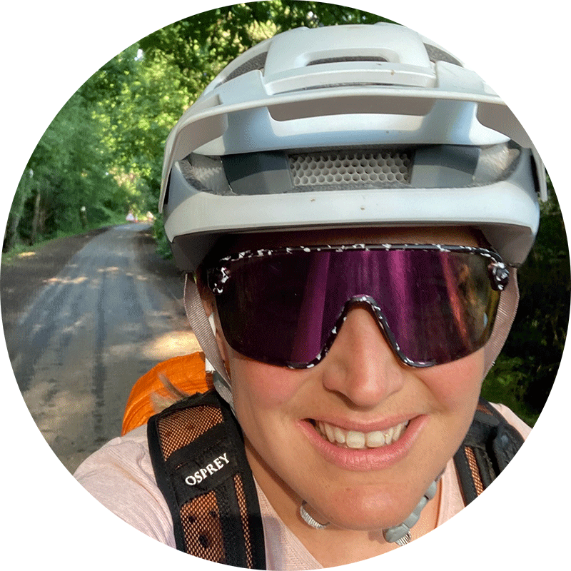 Rachael Walker in a white helmet and purple lens sunglasses on a tree-lined lane