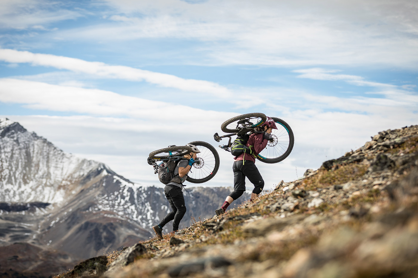 Two riders carry their bikes on their shoulders up a rocky alpine ridge, snowy peaks behind