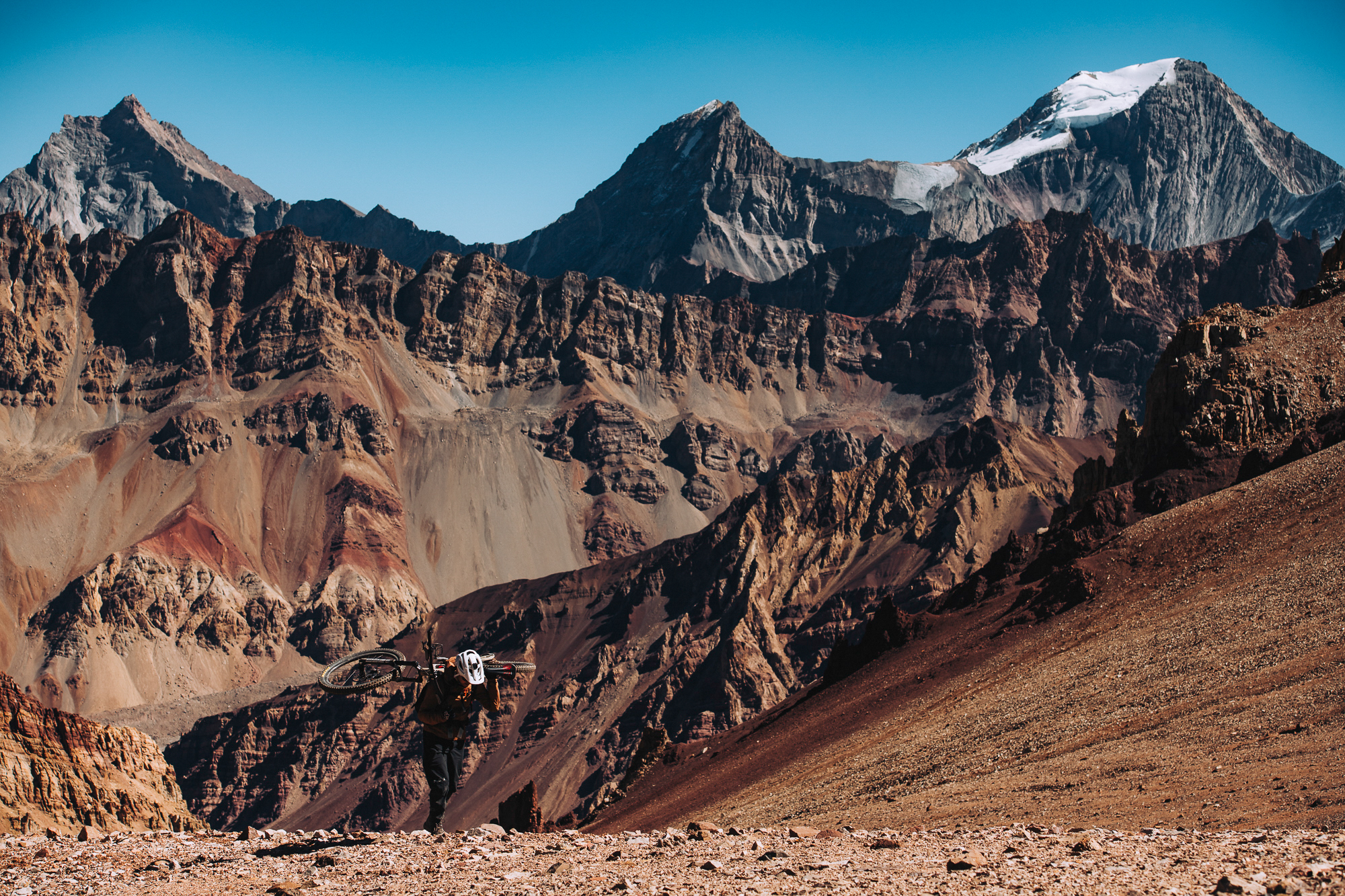 Rider carrying a bike below a huge banded rock face and snow-capped peak