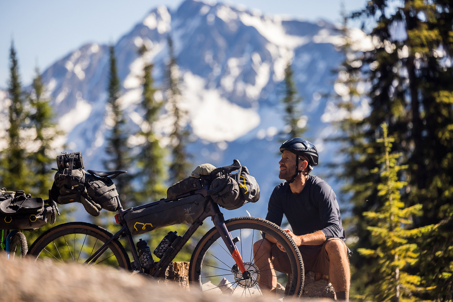 Rider resting on a rock beside a loaded gravel bike with a snowy peak behind