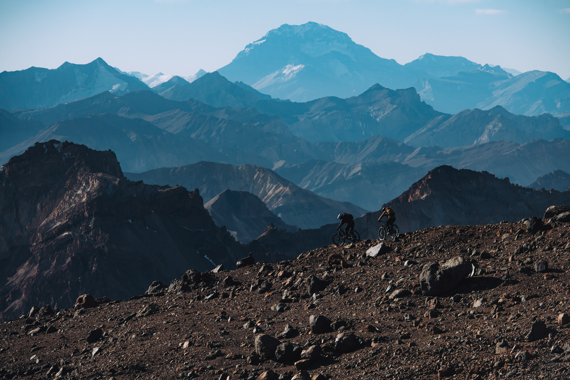 Two riders on a dark ridge with a huge snow-capped massif behind