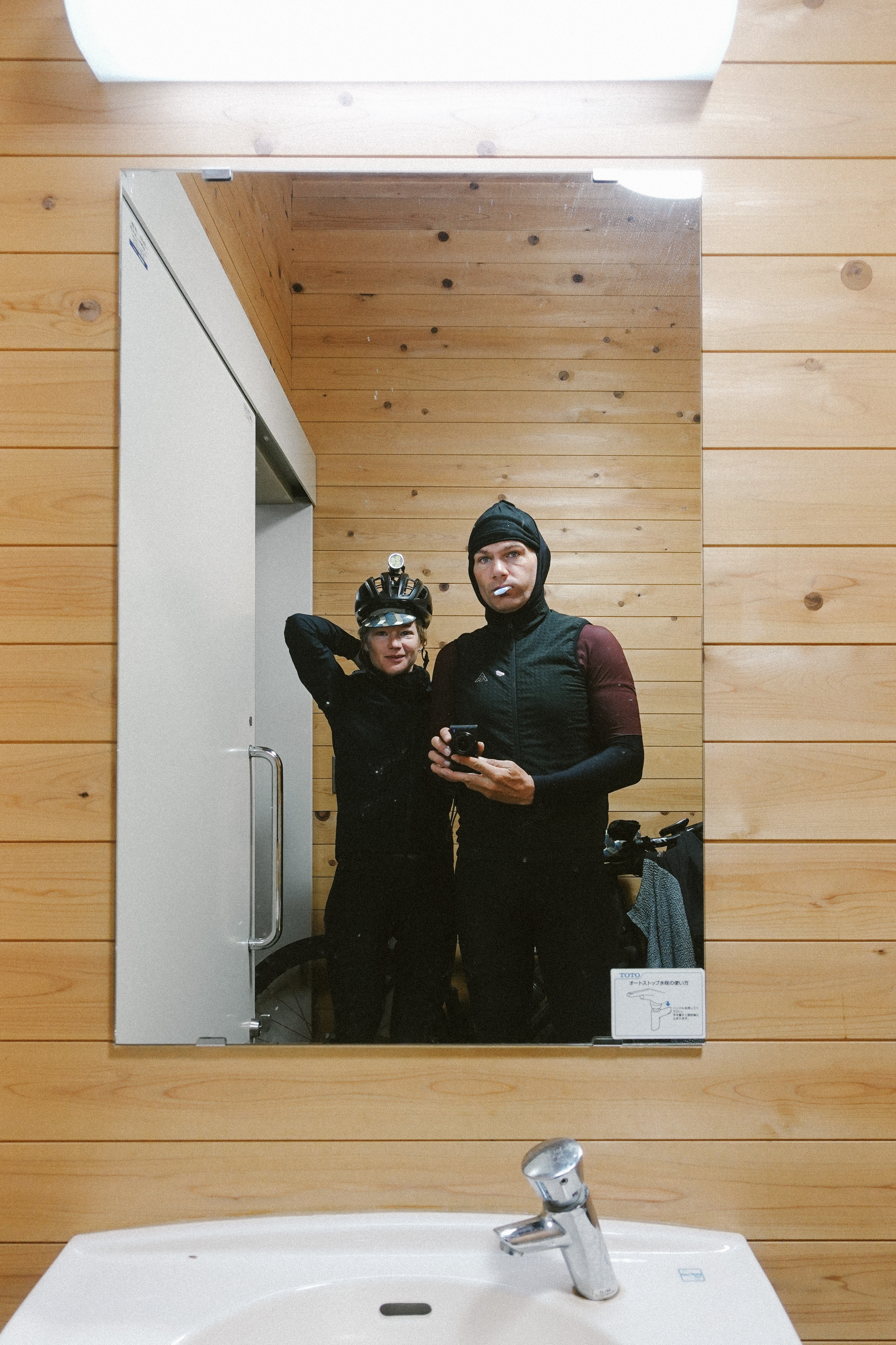 Two riders take a mirror selfie in a washroom, one brushing his teeth
