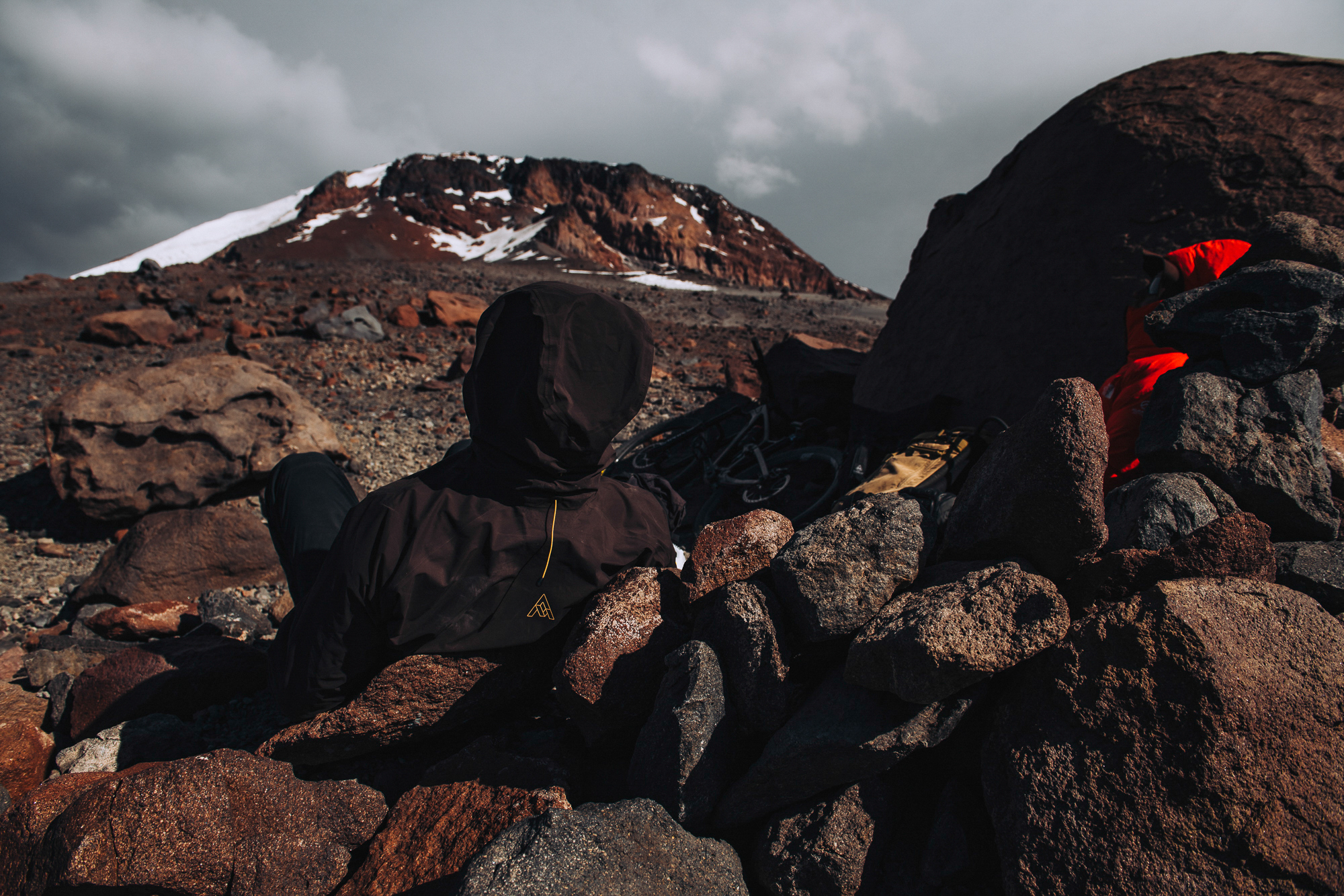 Rider resting behind a stone wind wall below a snow-streaked summit