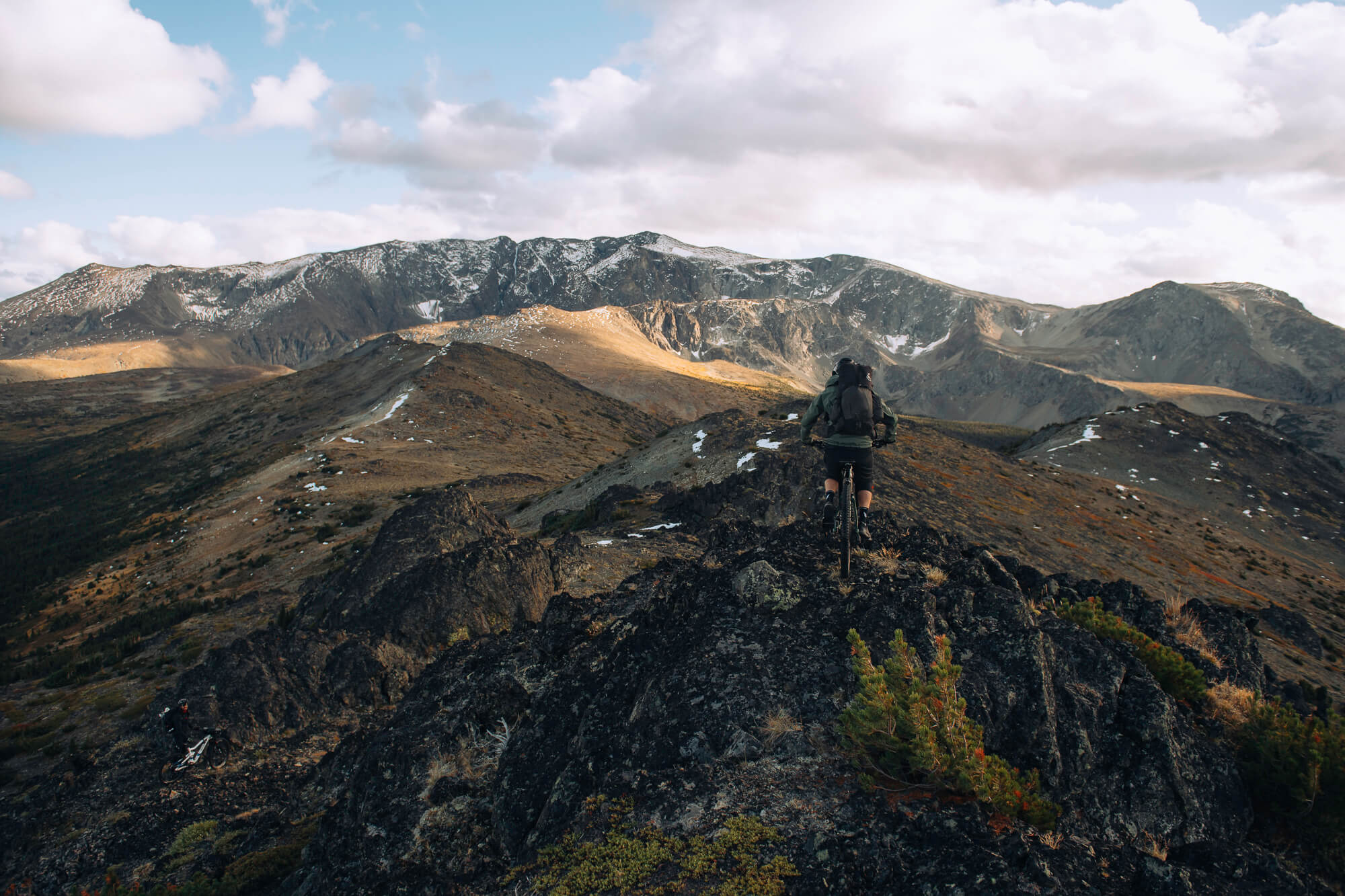 Rider on a rocky ridge crest facing a long snow-dusted mountain range