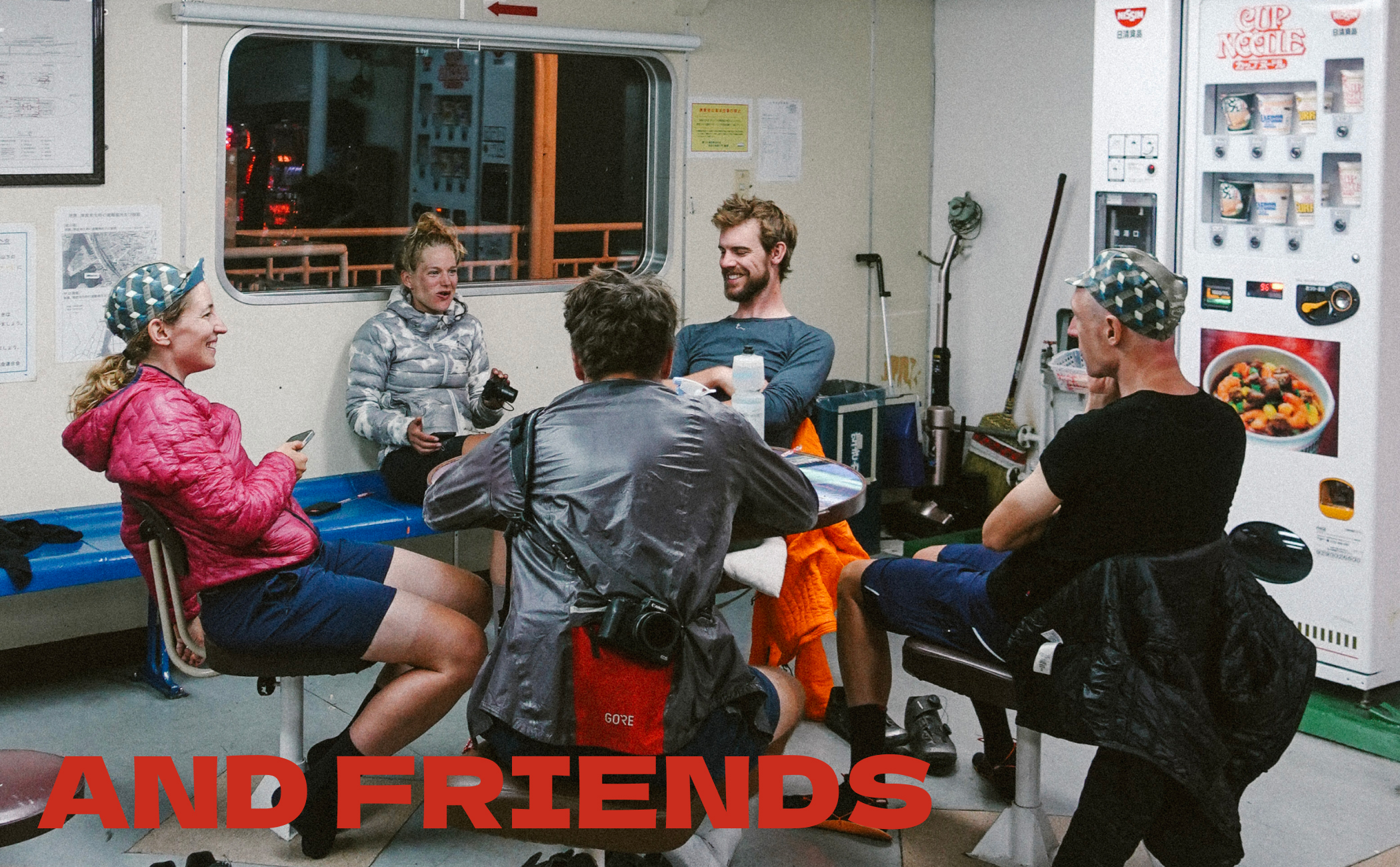 Riders around a table in a ferry lounge beside vending machines, captioned And Friends