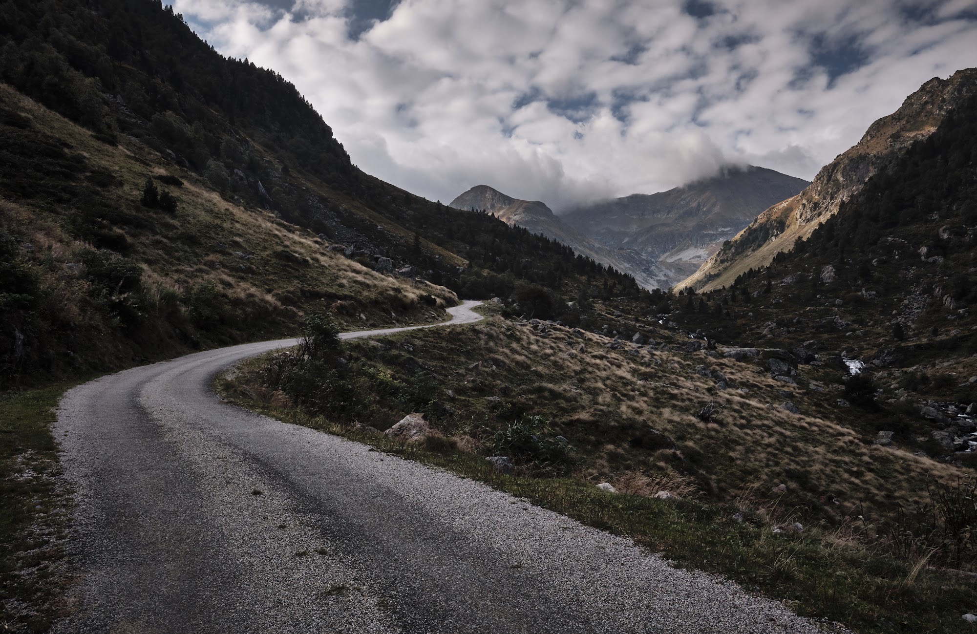 A narrow road winds down a steep mountain valley beside a stream under broken cloud