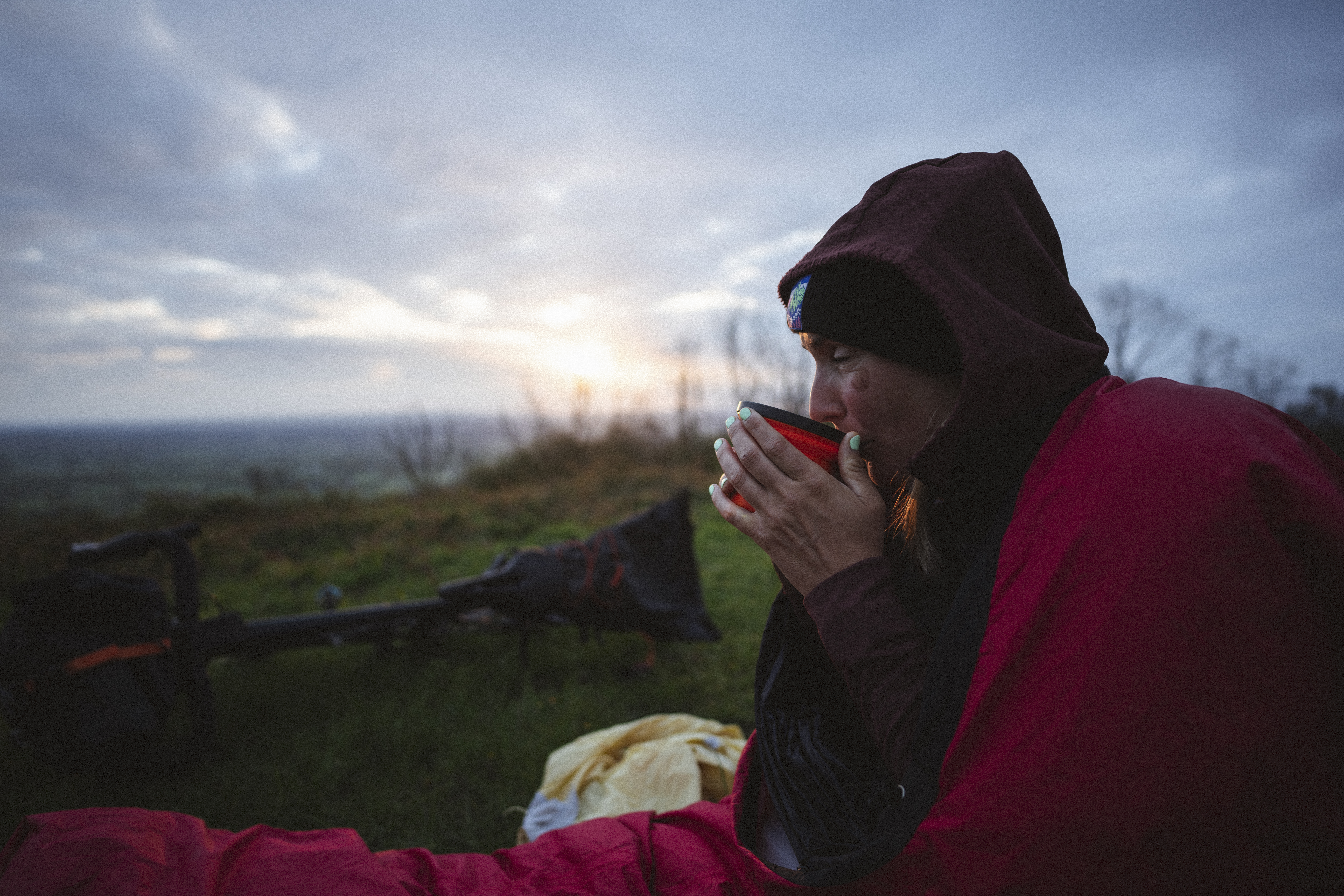 Rider in a hood drinking coffee from a red mug beside her bivvy