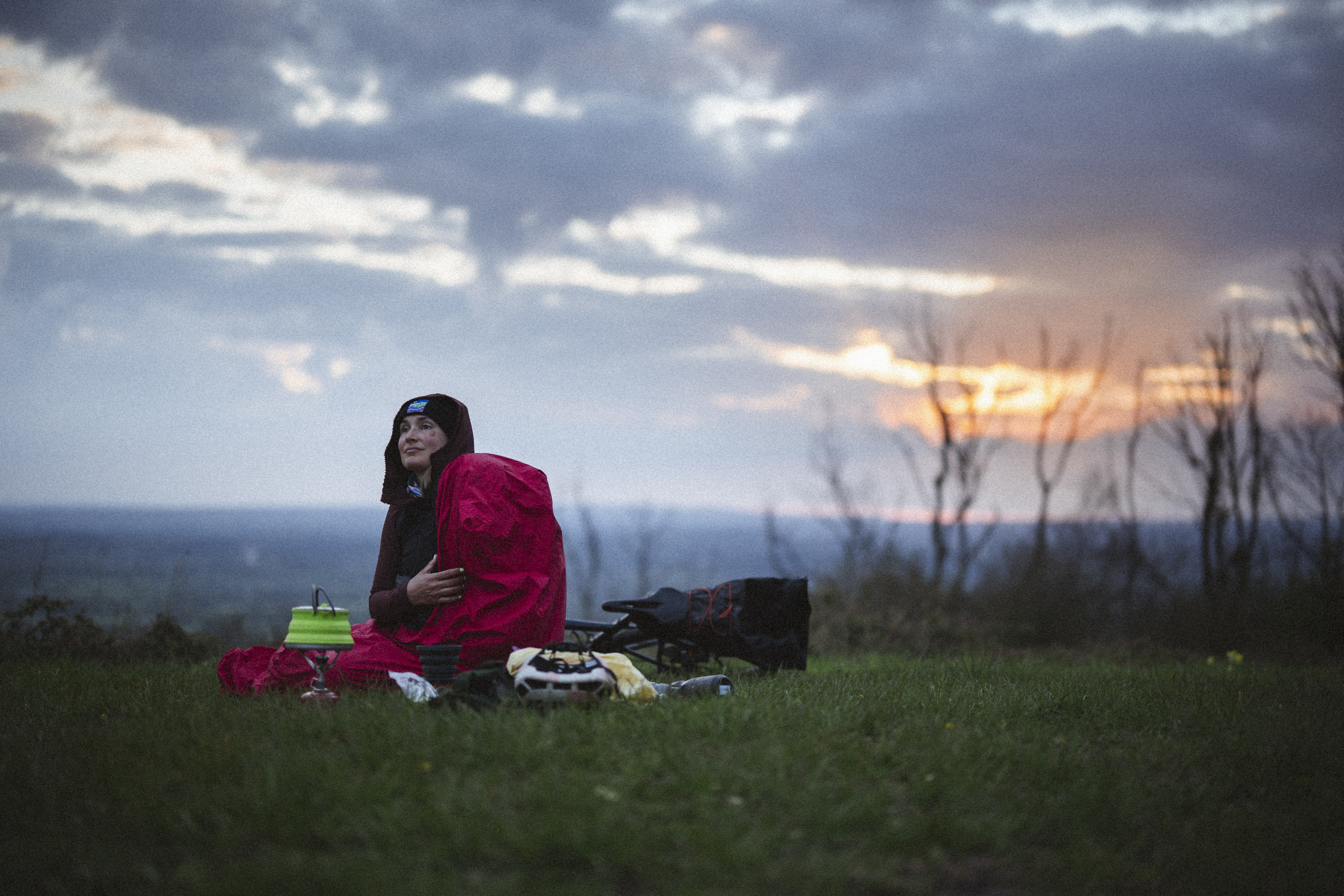 Rider sitting in a bivvy bag on a grass hilltop watching a cloudy sunrise