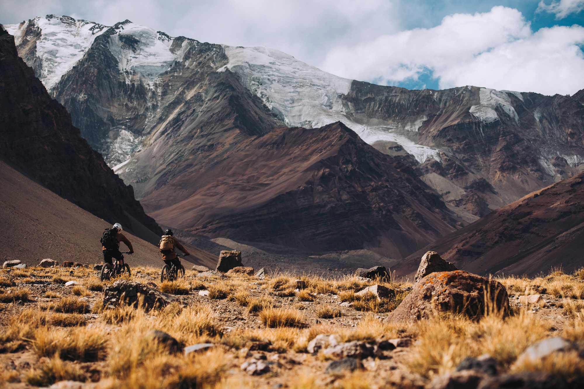 Two riders pushing bikes across yellow grassland below a hanging glacier