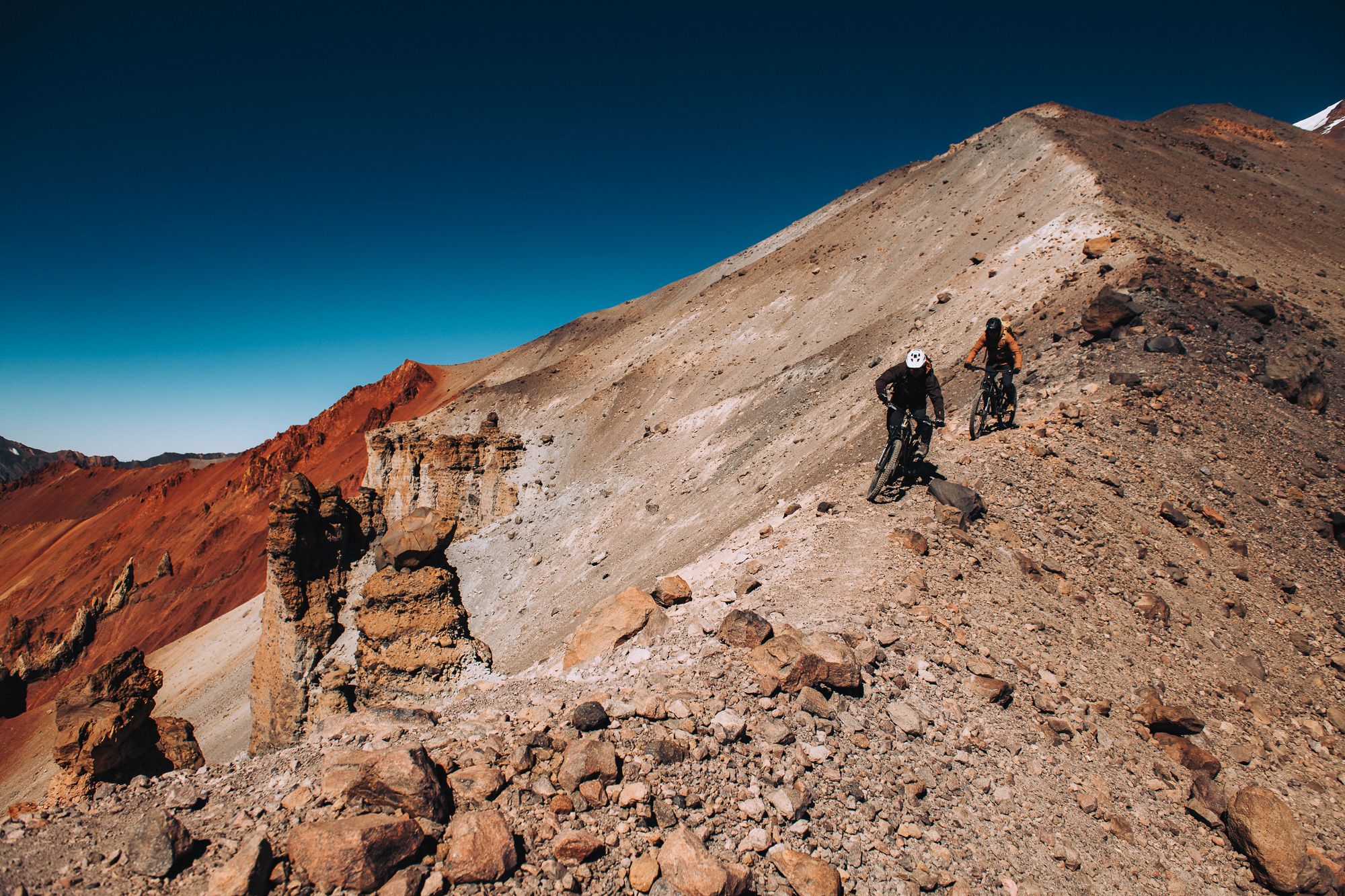 Two riders descending a broad ash-covered ridge under deep blue sky