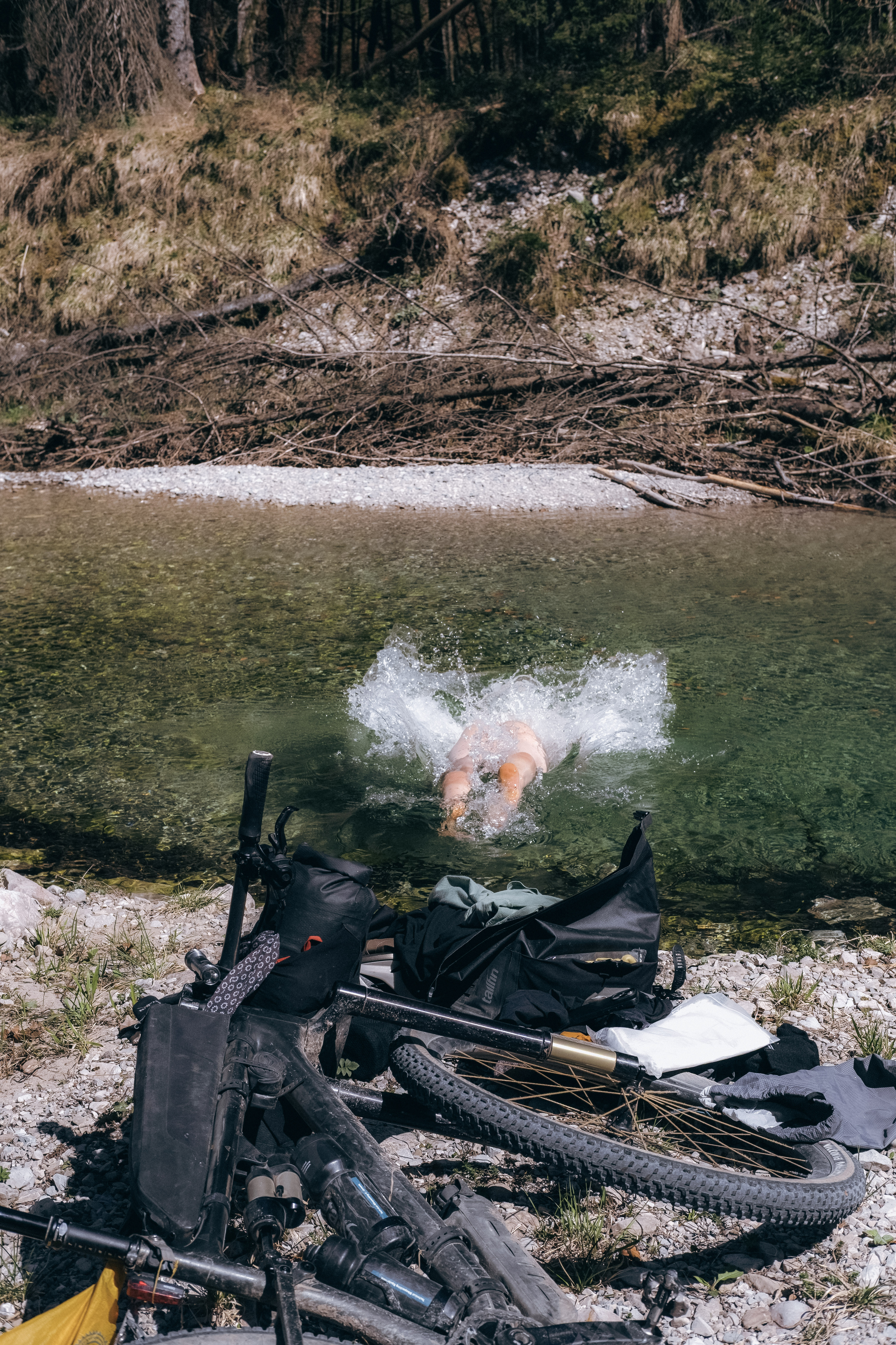 Swimmer diving into a clear green river beside loaded bikepacking bags on the stones