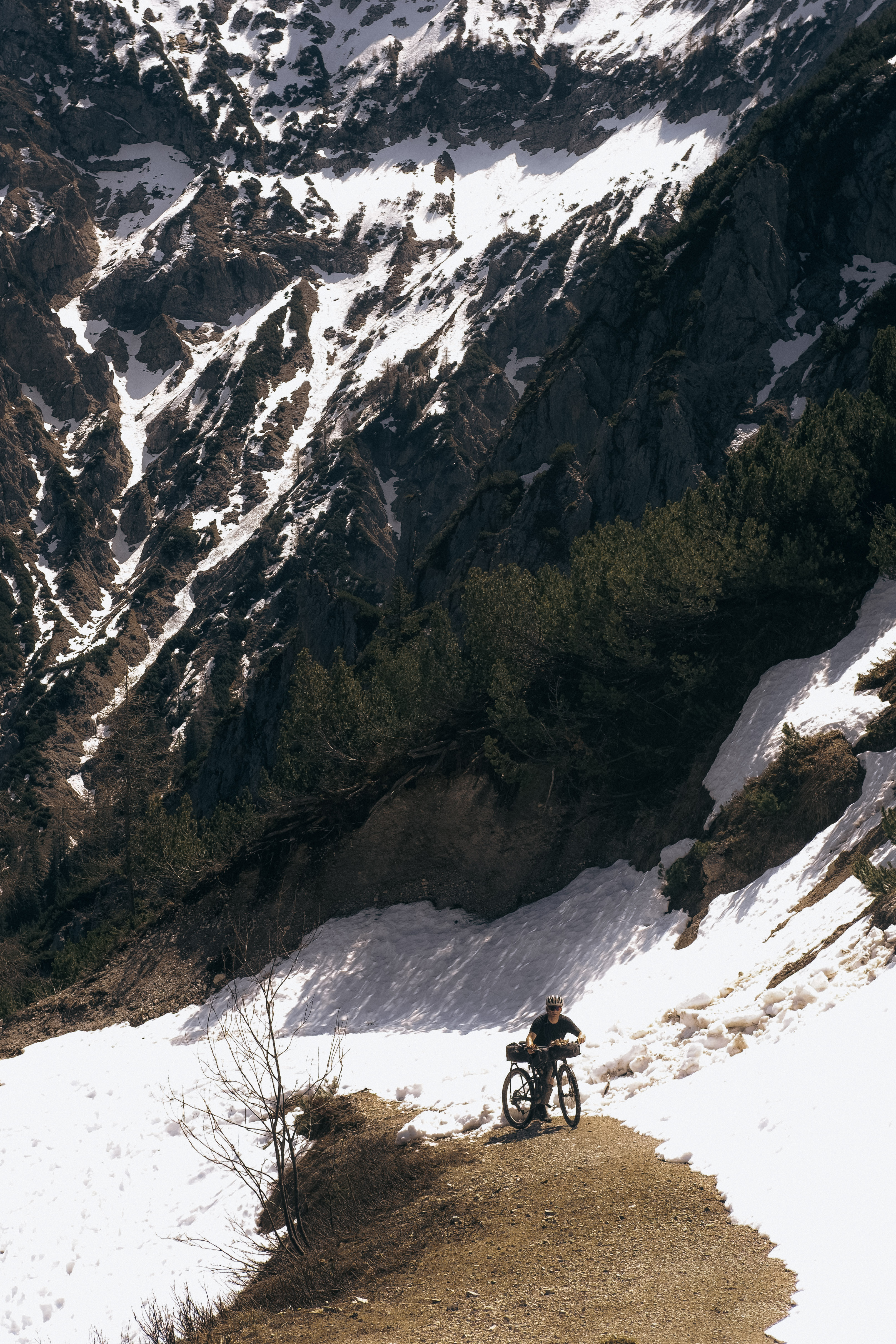 Rider pushing a loaded bike along a gravel track cut through deep spring snow