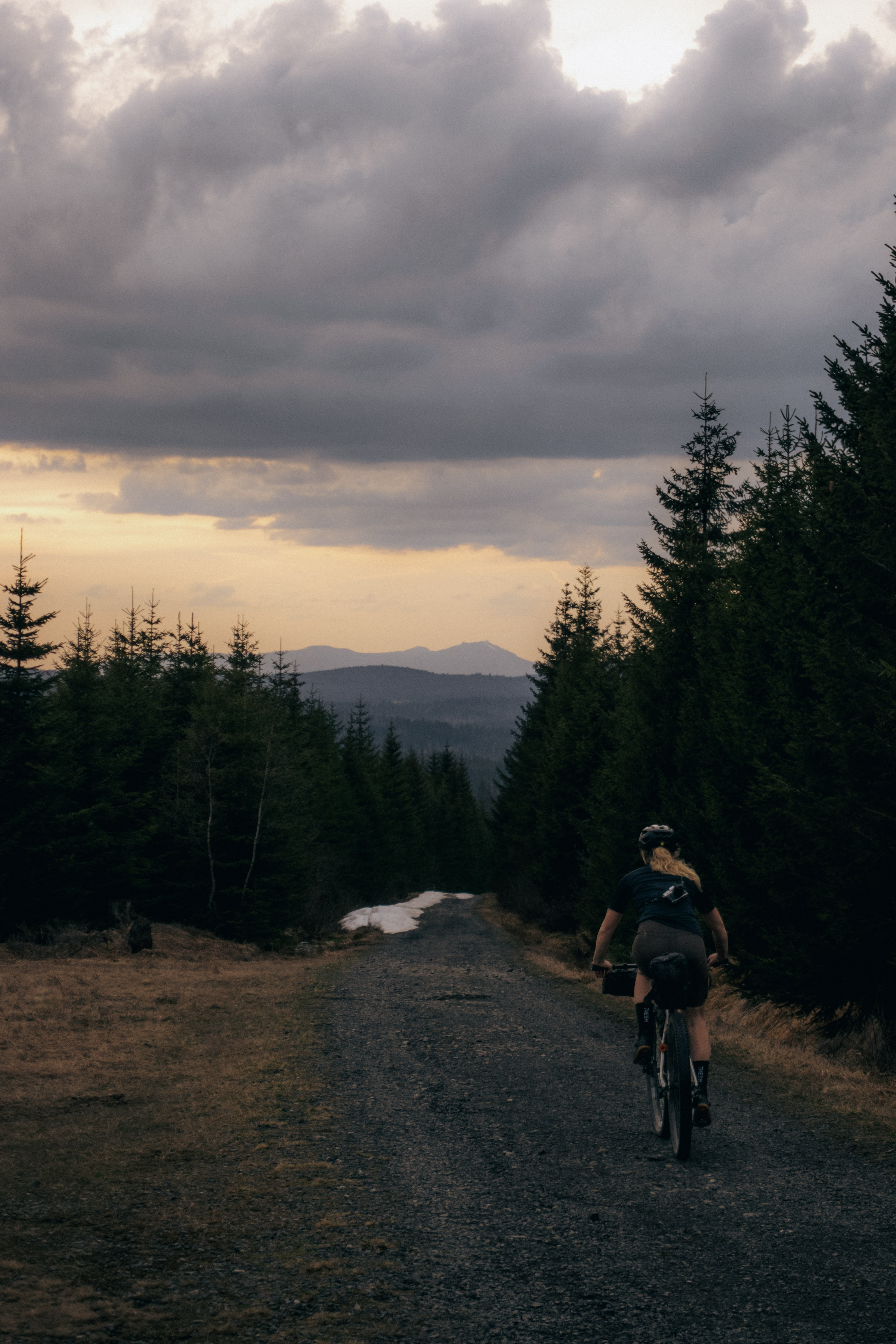 Rider on a gravel forest road at dusk, a distant peak on the horizon and snow beside the track