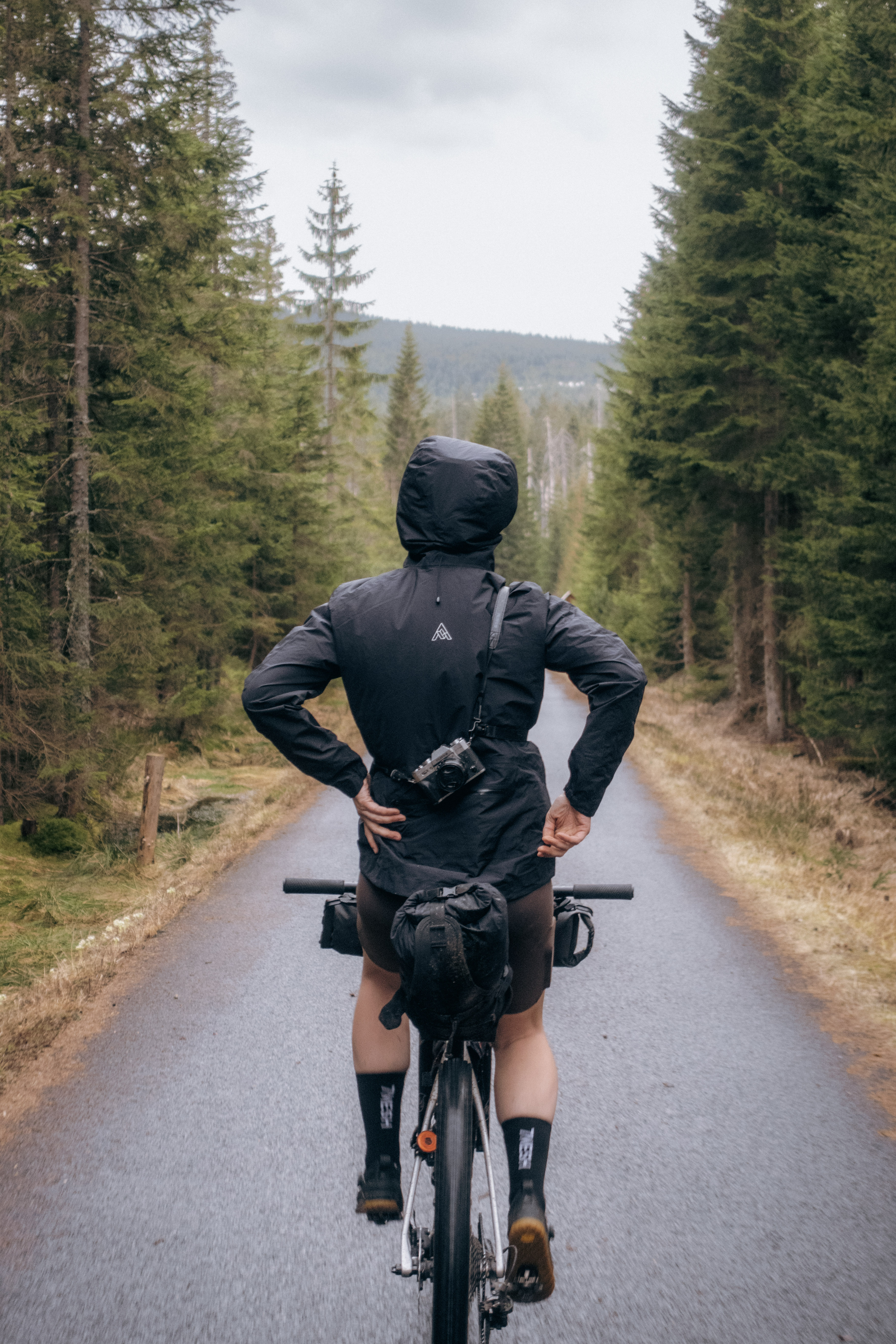 Rider from behind in a hooded black rain jacket, a film camera slung across their back