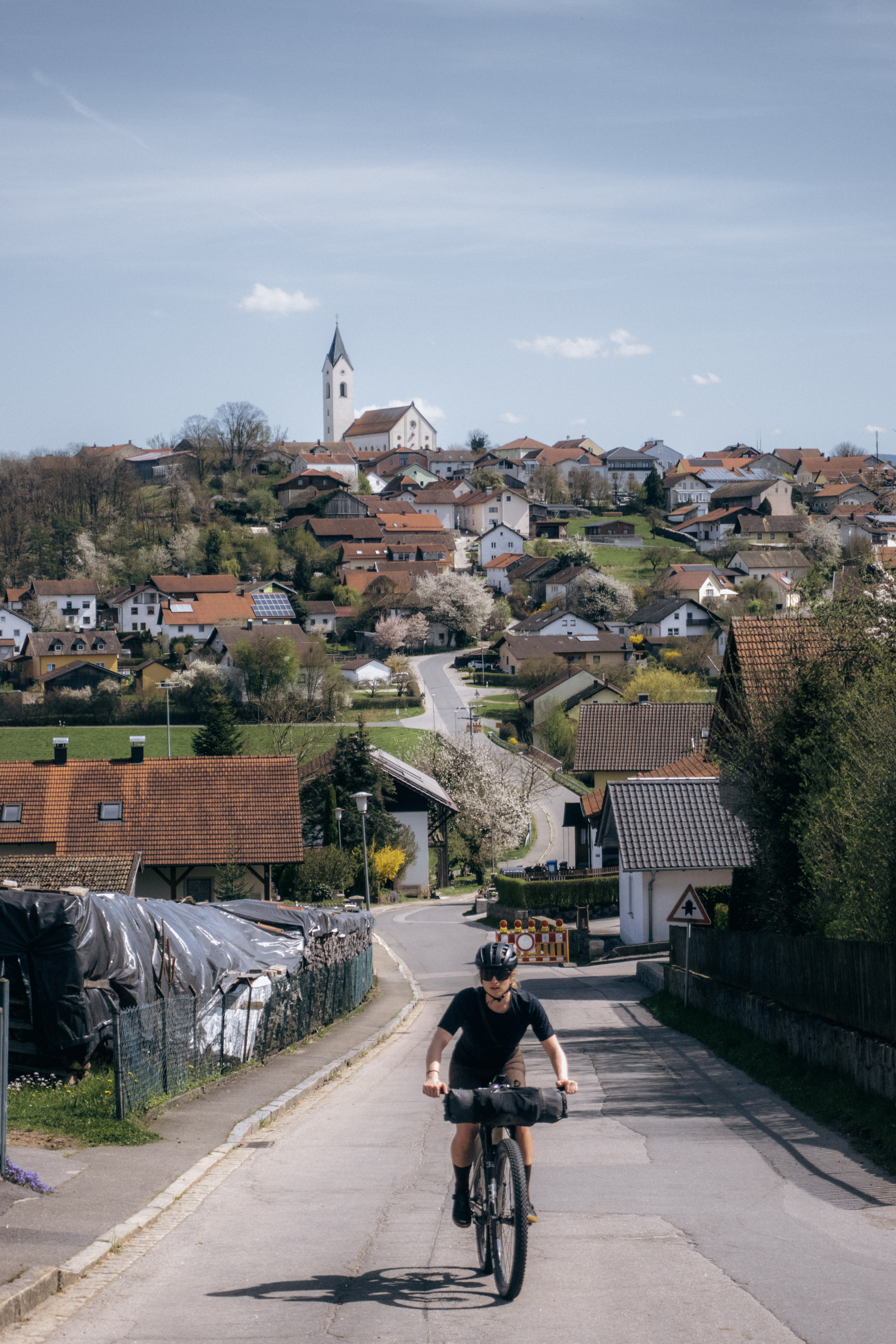 Rider on a loaded bike coming up a village street past houses and roadworks barriers